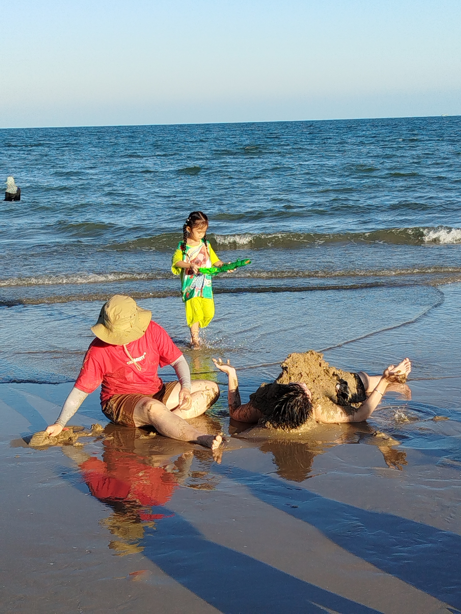 A father and child play on a beach. The father, wearing a red t-shirt, brown shorts, sun sleeves, and a sun hat, sits near the shore, partially reflected in the wet sand. Another person, covered in sand, lies on their back. A child wearing a yellow and green outfit stands in the water with a green toy. The scene is set on a beach near the ocean, under a clear sky. The scene may be near Ho Chi Minh City, Vietnam, but there is no indication on the image.