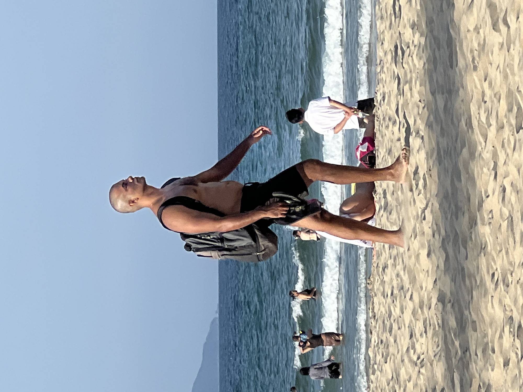 A bald man in black shorts and no shirt walks along a sandy beach carrying a backpack and holding a pair of flip-flops. He is facing right, looking out towards the ocean. Behind him, people are wading in the sea, and waves are breaking. In the background, mountains are visible in the distance. The scene is likely Đà Nẵng, Vietnam, given the presence of a beach and mountains.