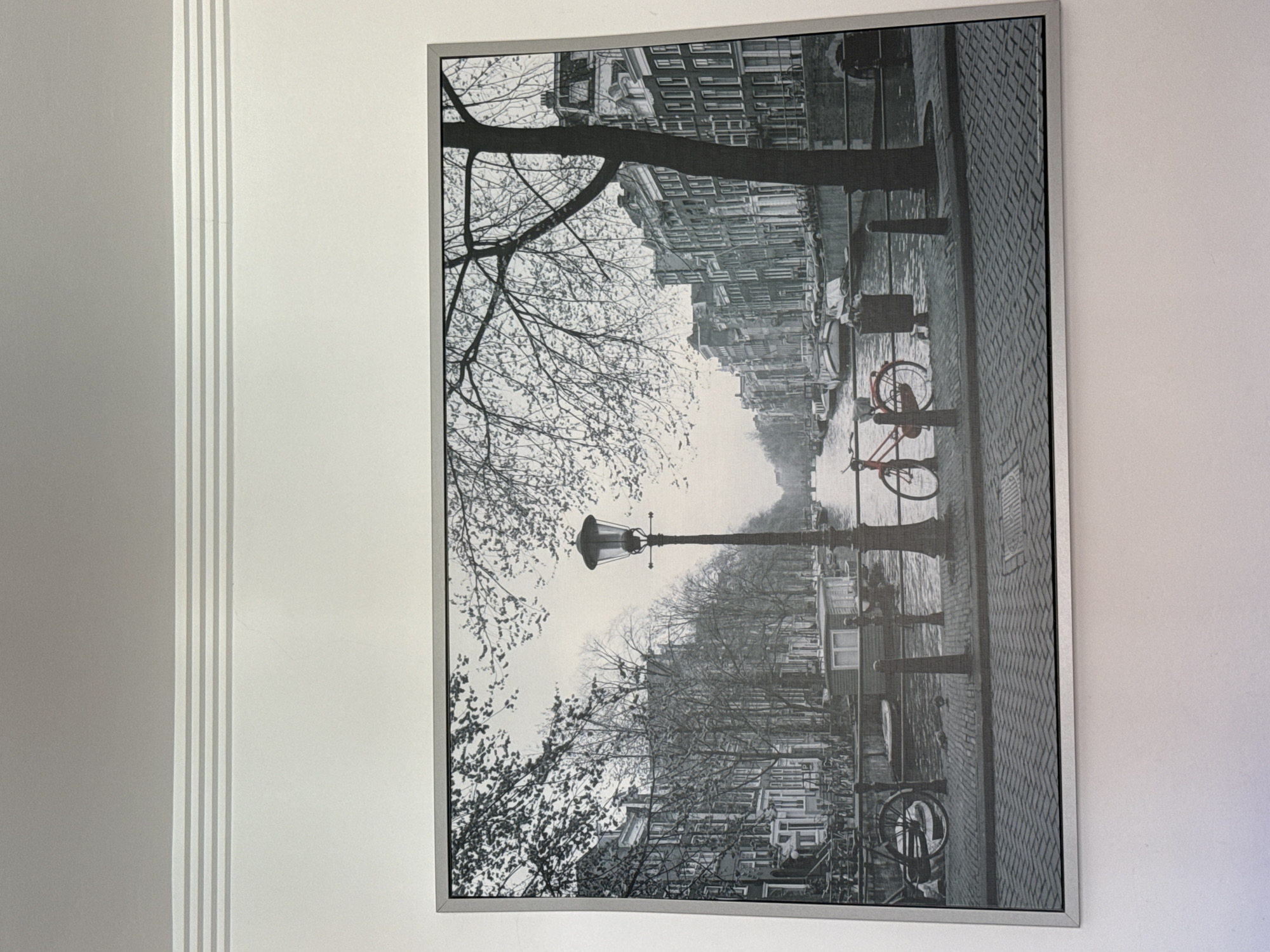 A framed black and white photograph depicts a picturesque canal scene in Amsterdam, Netherlands. The image showcases traditional Dutch architecture with narrow, tall buildings lining the canal. A vintage lamppost stands prominently on the cobblestone walkway, casting a warm glow. In the foreground, a red bicycle is parked, adding a splash of color to the monochrome scene. Several boats are moored along the canal, and trees with bare branches create a delicate canopy overhead. The overall atmosphere is serene and nostalgic, capturing the timeless beauty of Amsterdam. The location is not in Valencia, Spain.