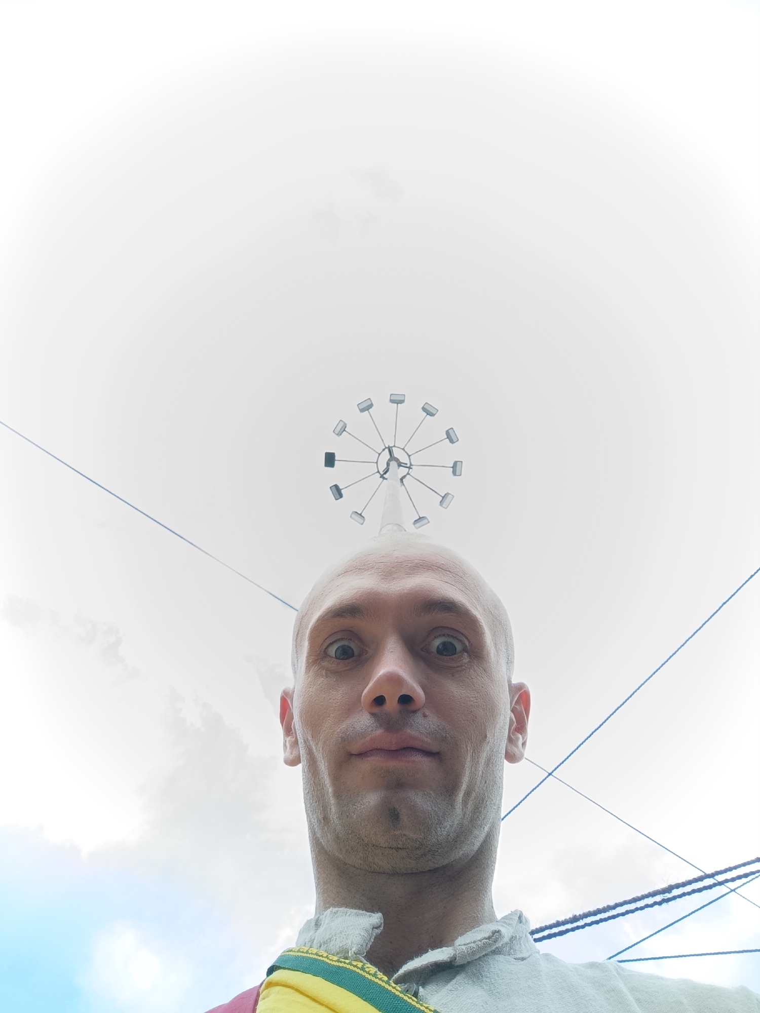 The image is a low-angle, wide-shot selfie of a bald, Caucasian man taken outdoors. He is wearing a light-colored garment with a yellow and green trim around the neck, and a hint of red fabric is visible on the left shoulder. His eyes are wide, and his mouth is slightly open, conveying a surprised or excited expression.
In the background, a white, overcast sky is visible, with some wisps of clouds. Suspended against the sky are several dark, thin wires or cables, creating a geometric pattern. Directly above the man's head and centered in the upper portion of the image is a Ferris wheel or a similar circular amusement park ride. The ride appears to be white with several rectangular pods or cabins attached around its circumference. The man's head and shoulders occupy the bottom half of the frame, with the Ferris wheel and sky filling the rest.
This image was taken in Manali, India.