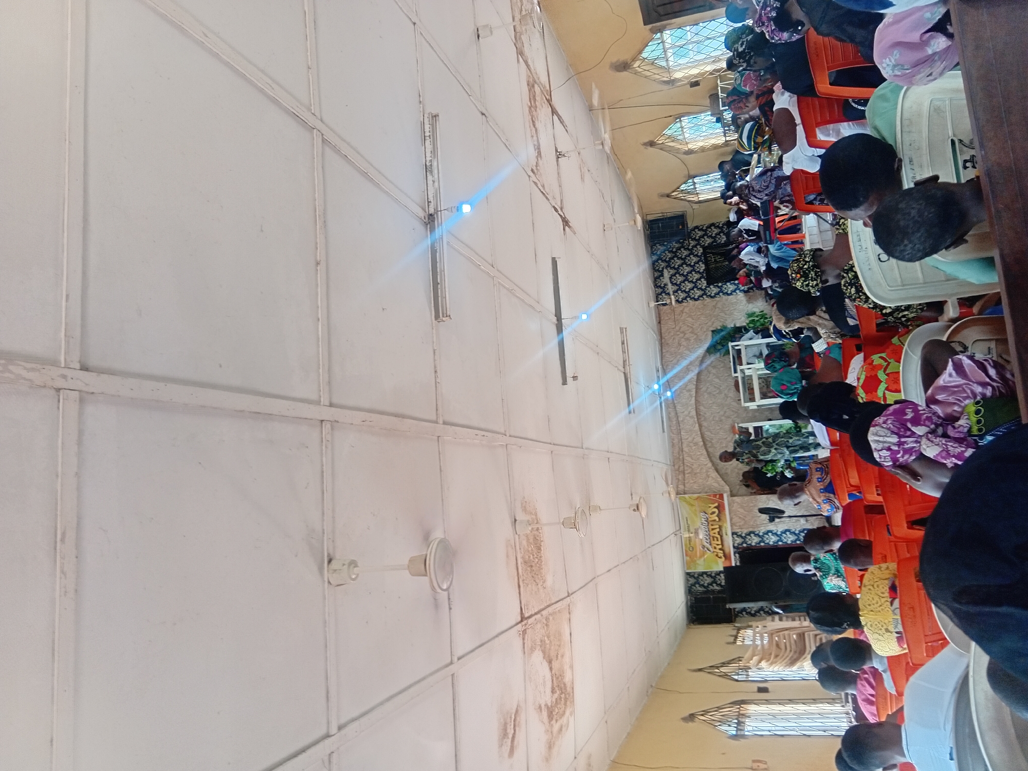 The image depicts an indoor gathering, likely a church service, taking place in the city of Ejule, Nigeria. The scene is viewed from behind a seated congregation, facing the front of the room where a speaker stands on a raised platform. The ceiling is a white, tiled surface with visible beams and several white ceiling fans. Some areas of the ceiling show water stains. Fluorescent lights and hanging bulbs illuminate the space.
In the foreground, individuals are seated on orange and white plastic chairs, their backs to the camera. Many are wearing colorful clothing and headwraps. The middle ground shows more seated attendees and a speaker, dressed in a patterned garment, addressing the audience from a stage decorated with fabric and a banner that reads "Exceeding Great Joy". There is audio equipment visible, including speakers and what appears to be a podium or lectern. Stacked chairs are visible to the left of the stage.
The walls of the room are painted a light yellow. Tall windows with barred panes are positioned along the side of the room, allowing natural light to enter. The overall impression is of a community gathering in a simple, functional religious space within Ejule.