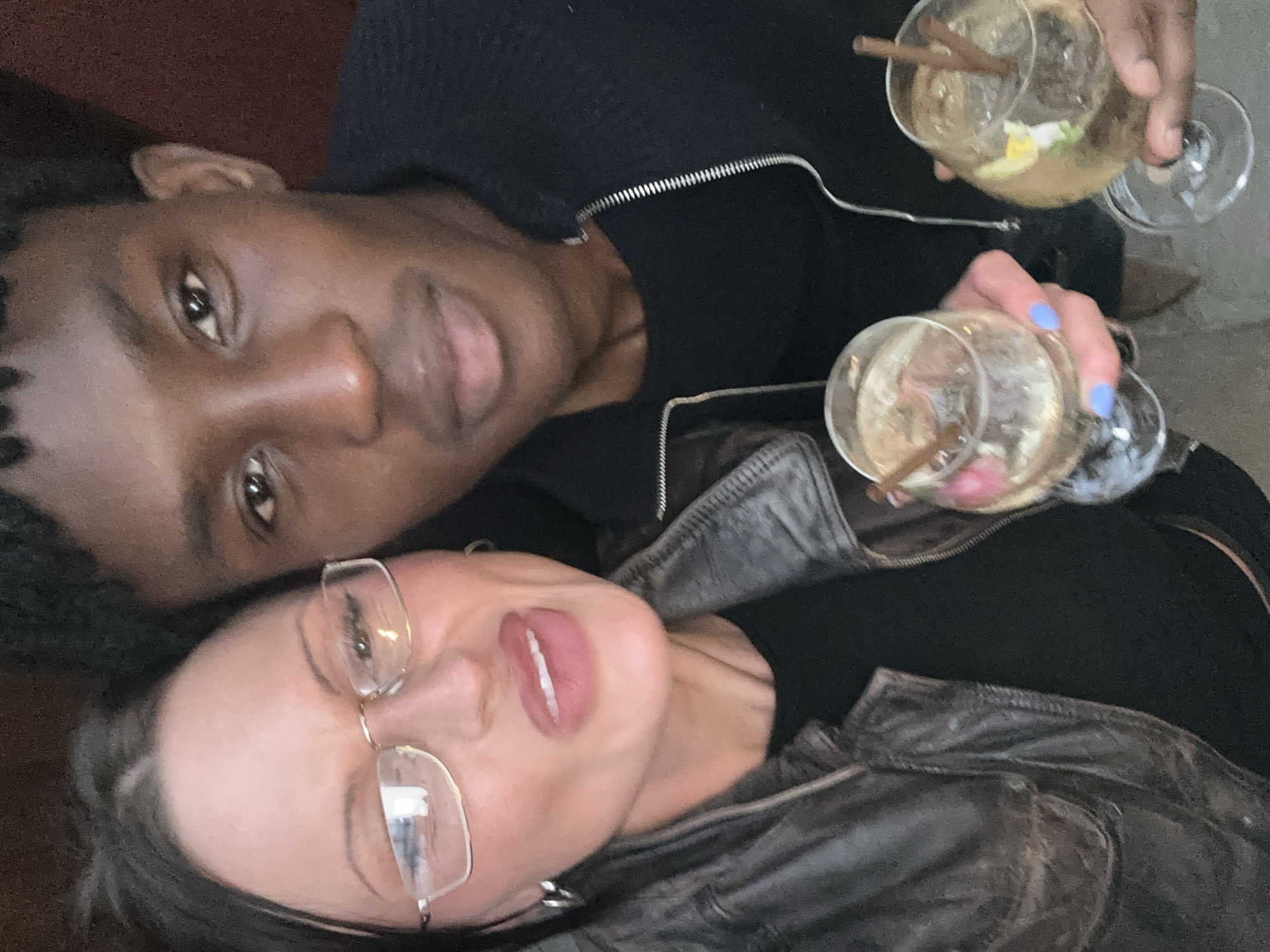 Two people are shown holding drinks. The person on the left has dark skin and short, curly black hair. They are wearing a black, zipped sweater. The person on the right has lighter skin and glasses. They are wearing a black shirt and a dark, distressed leather jacket. Both are holding glasses filled with a clear liquid, ice, and a cinnamon stick. The location is not identifiable from the image.