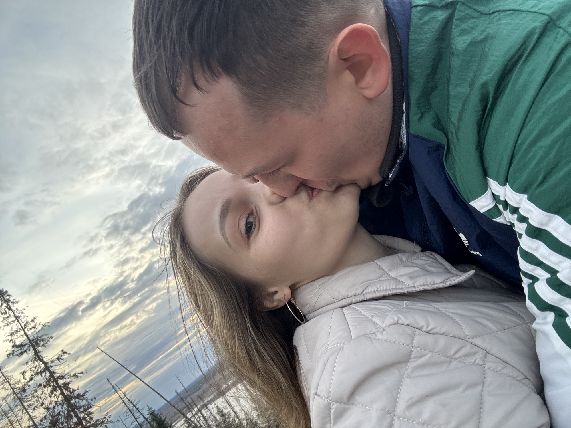 A man and woman are kissing outdoors. The woman is wearing a light-colored quilted jacket and gold hoop earrings. The man is wearing a green jacket with white stripes. The background shows trees and a cloudy sky. The photo appears to be taken in the city of Noviy, Russia.