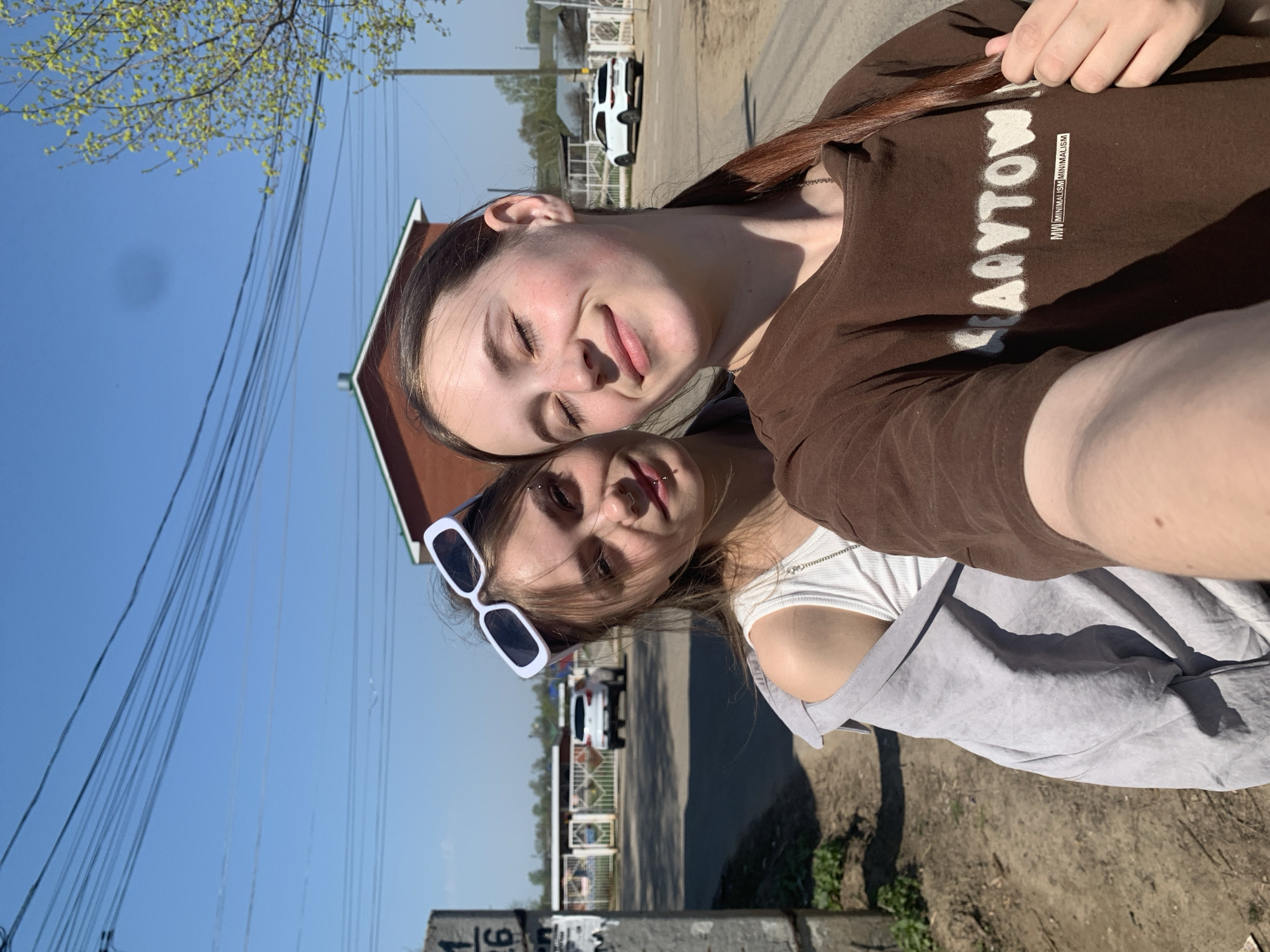 Two young women are closely positioned, likely in a selfie. The woman on top has brown hair, closed eyes, and wears a brown t-shirt with "LARYTOW" printed on it. The woman below wears white sunglasses, a white top, and a grey overshirt. They are in Фоки, Russia. A street scene is visible in the background with a building and cars. There are utility poles and wires overhead. A concrete structure is in the foreground with obscured markings. The weather appears sunny.