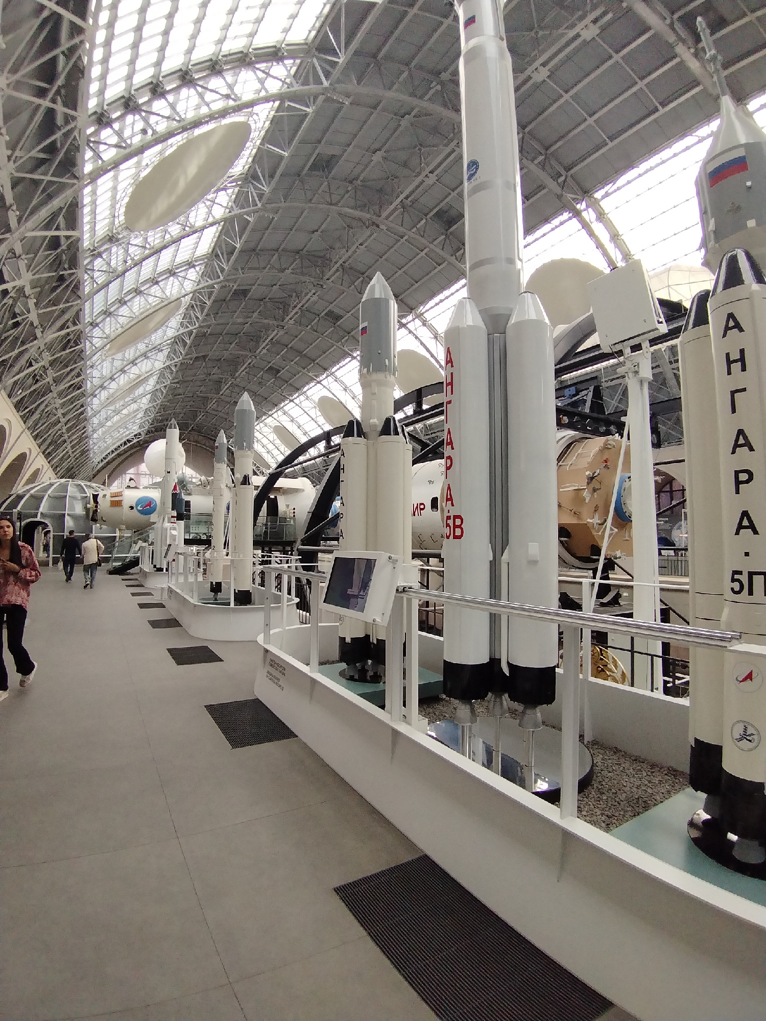 The image depicts the interior of a museum exhibit, likely the Cosmonautics Memorial Museum in Moscow, Russia, given the prominent display of rockets and space-related artifacts. Several tall, cylindrical rocket models are arranged in a line, with the closest one being the largest and most detailed. This rocket has white sections with black bases and appears to have Cyrillic lettering on its side, possibly indicating its name or model. Other, smaller rocket models are positioned behind it, along with a larger, capsule-like structure with windows. The museum's ceiling is a vast, complex structure of metal beams and translucent panels, allowing natural light to flood the space. In the foreground, a woman wearing a patterned shirt and dark pants walks towards the left of the frame. Two men are seen walking away from the camera in the background, further down the exhibit hall. The floor is a light-colored, tiled surface with black mats placed at intervals. The overall impression is of a well-lit, spacious museum dedicated to space exploration.