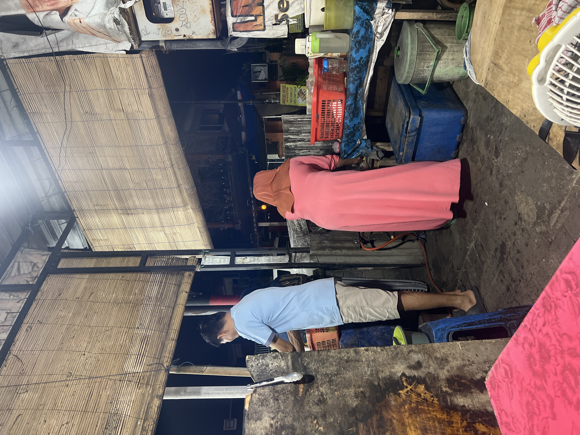 The image depicts an outdoor food stall scene at night in Gianyar, Indonesia. A man wearing a light blue t-shirt and beige shorts stands facing left, seemingly engaged in food preparation, with tongs held in his right hand. To his left, a portion of a wooden counter is visible. In the foreground, a bright pink surface with a floral pattern is present. A woman in a pink long-sleeved dress and a brown hijab stands facing away from the camera, to the right of the man.
Behind the man and woman, a table is set up with various items. A red plastic basket, a glass with liquid, and a green plastic container are on display. Blue and green ice chests are visible in the background, along with a green trash bin. The stall is covered with bamboo blinds, suggesting a rustic and open-air setup. The ground is paved with dark, somewhat uneven material, indicating a typical street-side environment. The lighting suggests it is nighttime, with artificial light illuminating the scene. A sign with text and graphics is partially visible on the right side of the image.
