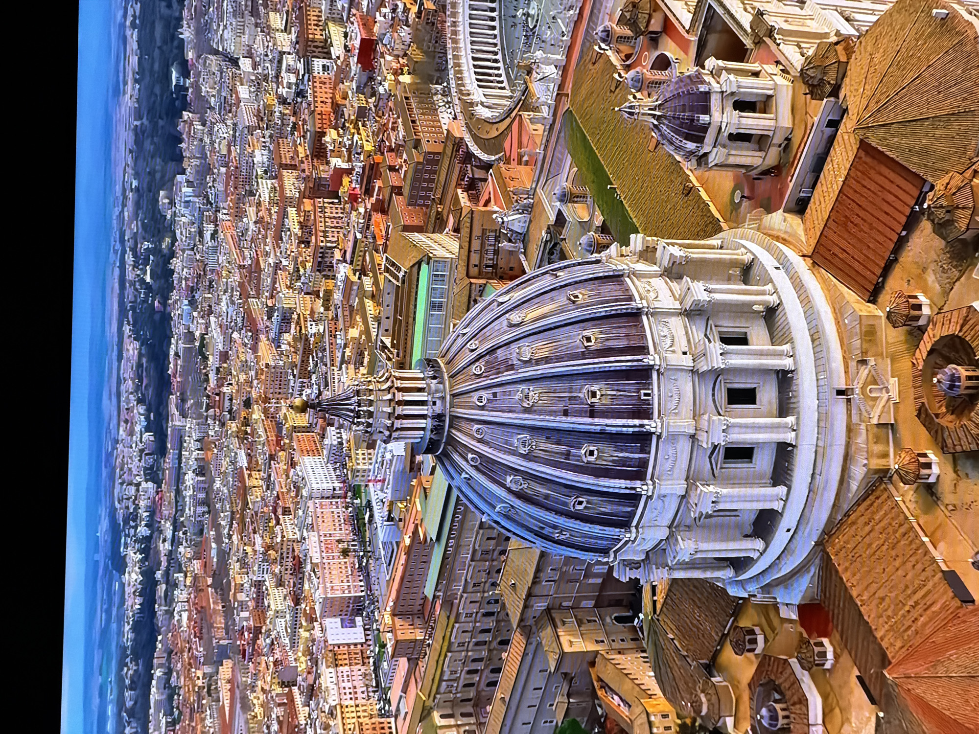 The image is an aerial view of St. Peter's Basilica and St. Peter's Square in Vatican City, Rome, Italy, with the city of Rome stretching out in the background. The prominent feature in the foreground is the massive dome of St. Peter's Basilica, which is intricately detailed with architectural elements. Surrounding the basilica are various buildings with terracotta roofs, characteristic of Roman architecture. In the mid-ground, St. Peter's Square, with its colonnades and obelisk, is visible, as are numerous other buildings in the city of Rome, displaying a dense urban landscape. The background shows a hazy horizon with distant hills. There are no people or animals clearly identifiable in the image due to the high altitude. The scene depicts a sunny day with clear skies, casting shadows on the buildings. This image is of Rome, Italy, not Chirchik, Uzbekistan.