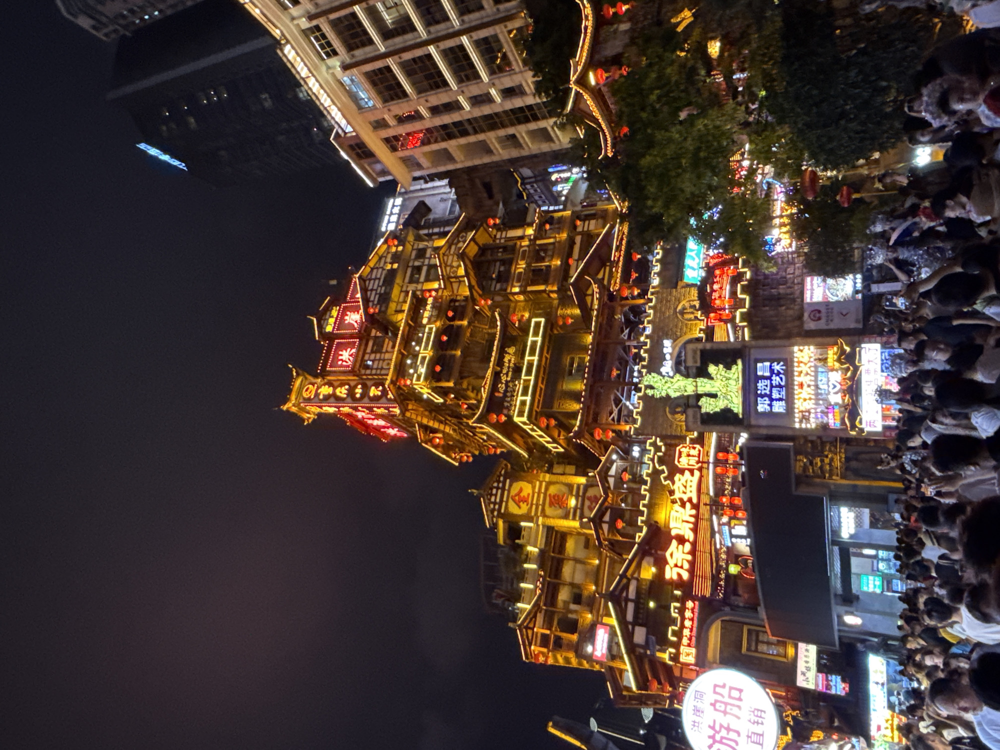 This is a nighttime, low-angle shot of the Hongyadong complex in Yuzhong District, China. The iconic, multi-story building, illuminated by warm yellow and red lights, dominates the frame. Its traditional Chinese architectural style, with layered roofs and intricate detailing, is accentuated by numerous red lanterns and neon signs. The building is surrounded by other structures, including a modern skyscraper in the background. A dense crowd of people is gathered in the foreground, many holding up their phones to take pictures. The atmosphere appears lively and festive, with the glowing lights and bustling activity suggesting a popular tourist destination.