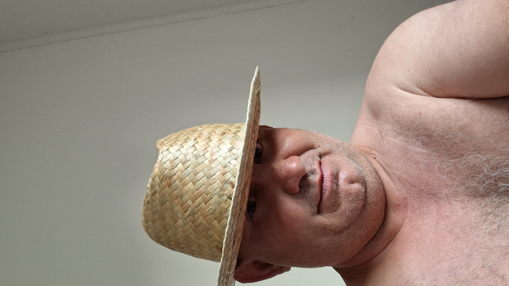This media file is a close-up, low-angle selfie of a person wearing a straw hat. The subject is a man with fair skin, a round face, and visible stubble. He is looking directly at the camera with a neutral expression. The straw hat is positioned to partially obscure his face, with the brim casting a shadow over his eyes. The background is a plain, light-colored wall, suggesting an indoor setting. The man's bare shoulder and upper chest are visible, showing light body hair. The image is oriented upside down. There is no visible text, and no specific cues about the time of day or weather are apparent. The context provided indicates the location is Hîncești, Moldova.