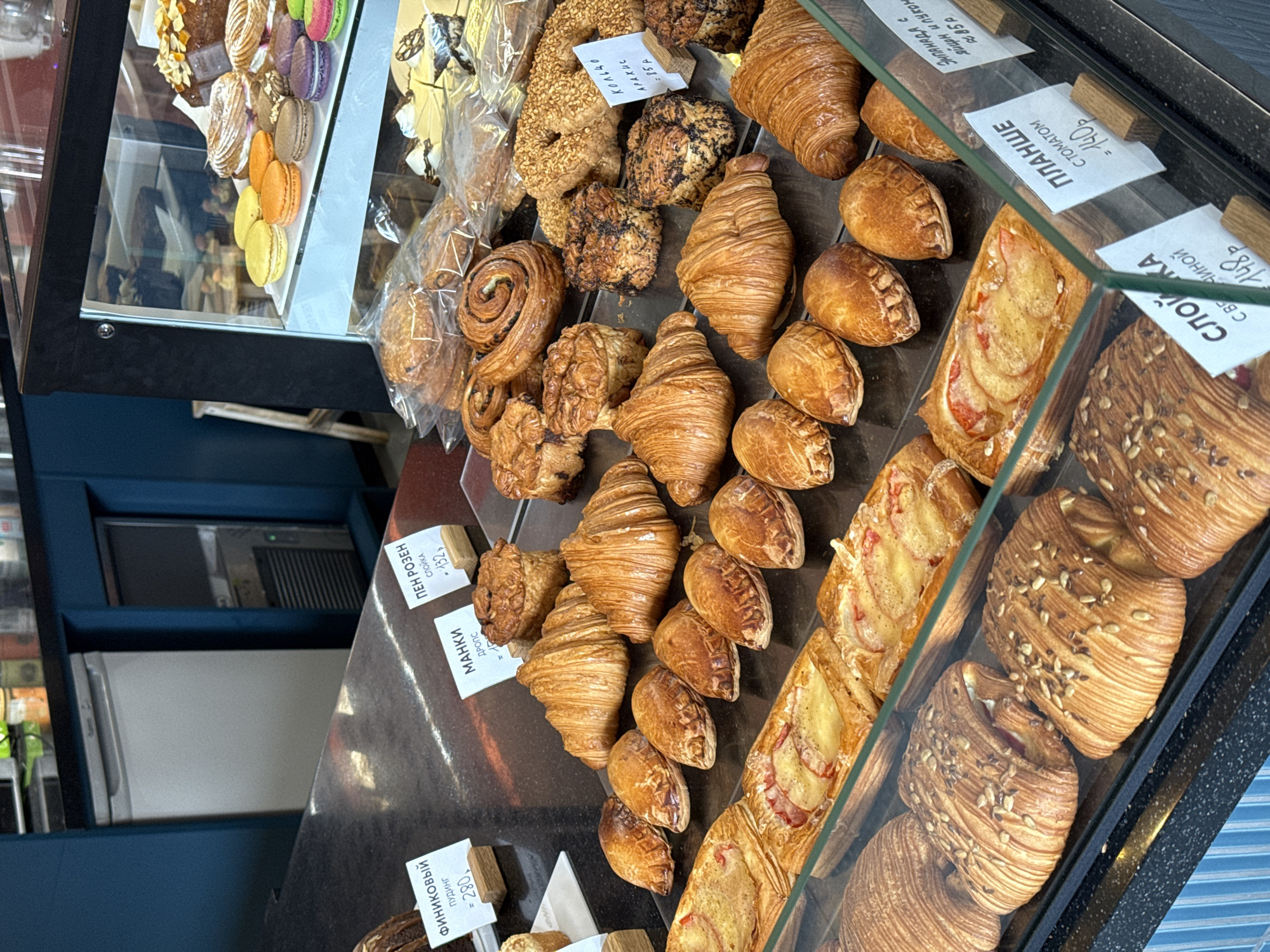 The image shows a display case of pastries in what appears to be a bakery or cafe. Various types of pastries are displayed, including croissants, cinnamon rolls, and fruit-filled pastries. Price tags in Russian are visible next to some of the pastries. The background shows the interior of the cafe, including a refrigerated unit. The pastries are displayed in rows on a dark surface. The image was taken in Krasnoyarsk, Russia.