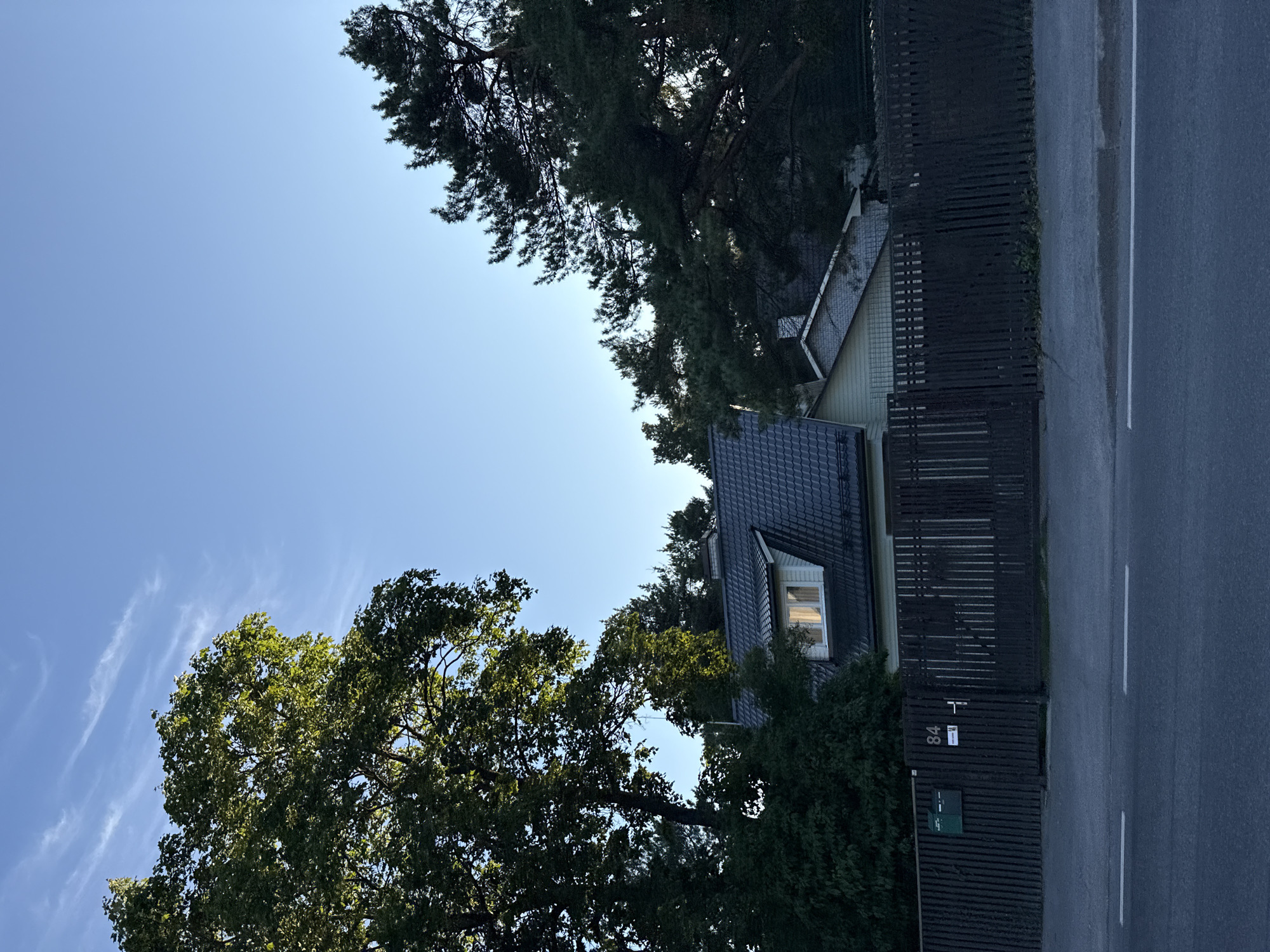 The image shows a residential property located at 84 Tihase Street in the Kristiine district of Tallinn, Estonia. The property features a house with a dark grey tiled roof and light-colored siding, visible through a dark wooden fence. A large tree with green foliage is situated to the left of the house, and a pine tree is on the right. A green mailbox is attached to the fence, and a house number "84" is also displayed on the fence next to the gate. The scene is captured from a street-level perspective, with a paved road in the foreground. The sky is clear blue with some thin white clouds, indicating it is daytime.