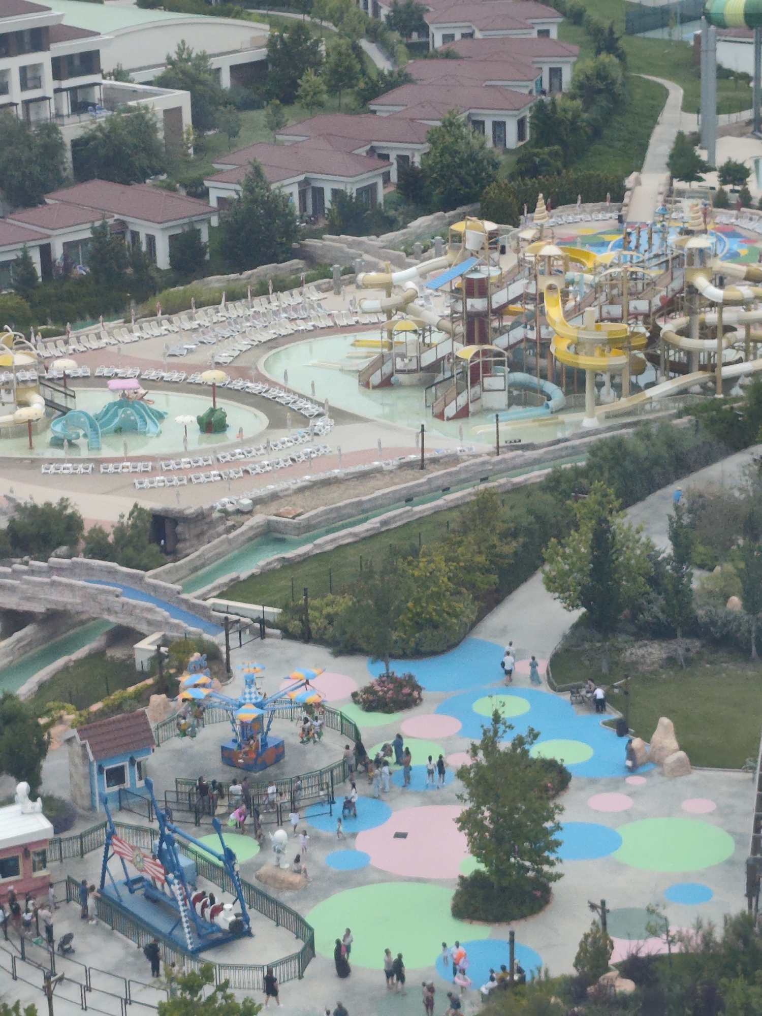 An aerial view captures a water park in Aktau, Kazakhstan, characterized by a complex of vibrant slides, pools, and recreational areas. The scene is divided between an organized water park with numerous colorful slides and a resort complex with rows of white lounge chairs bordering a large pool. In the background, a modern resort building with a white facade and dark windows stands prominently, surrounded by lush green trees. The water park itself features a variety of water slides, including a large yellow spiral slide and a smaller blue slide, nestled around a central pool area. Children and adults are seen scattered throughout the park, some engaging in water activities, while others relax on lounge chairs. The water park also includes a smaller area with a carousel and a swing ride, where children can be seen enjoying themselves. The overall environment is one of leisure and recreation, with the water park and resort forming a cohesive entertainment destination.