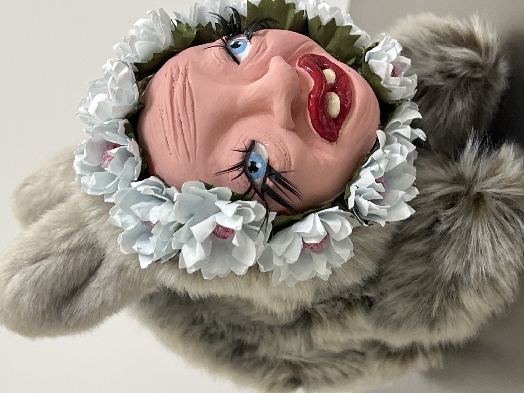 A whimsical, handcrafted doll's head lies within a fluffy, grey stuffed animal. The doll's head is made of a pinkish material, possibly clay or polymer, with exaggerated features including large blue eyes with thick, black lashes, and a large, open red mouth with white teeth. The head is encircled by a floral wreath of light blue and white flowers with pink centers and green leaves. The stuffed animal appears to be made of long-haired, grey-toned faux fur.
The image appears to be staged, with the object positioned against a white or off-white background.
The image's location in the city of Saint Petersburg, Russia, cannot be determined based solely on the visual information provided. There are no landmarks or indicators to reveal the location.