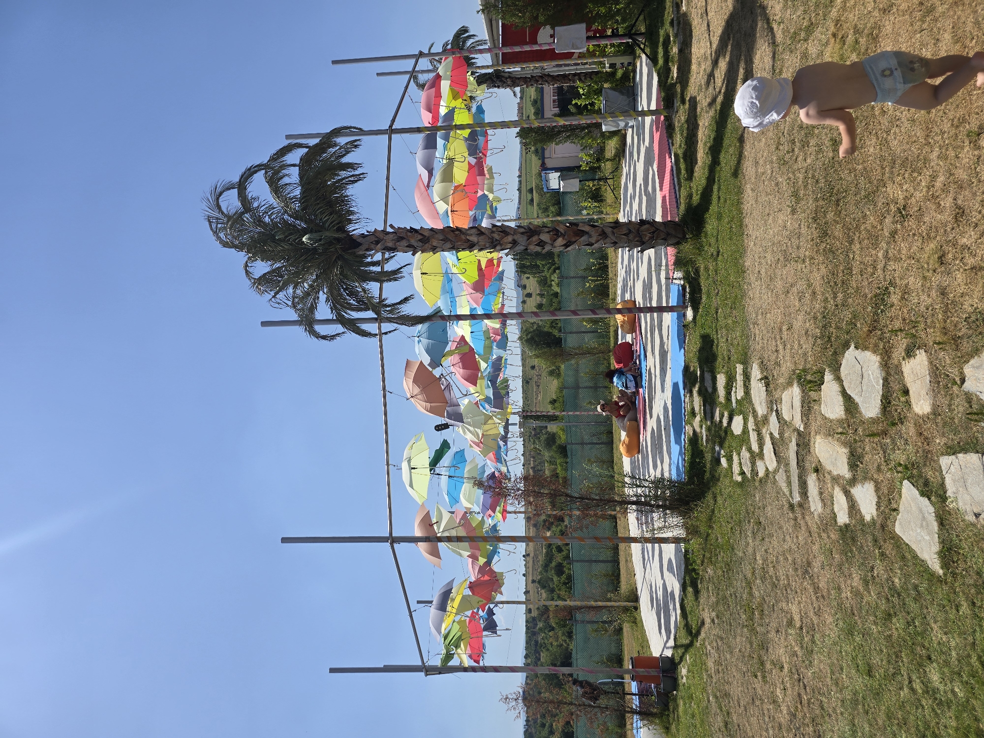 In an outdoor, sunny setting in Unknown, Unknown, a fenced-off area features a circular stage adorned with numerous colorful umbrellas suspended overhead. A palm tree stands centrally behind the stage. Several people are seated on colorful cushions on the stage. A young child wearing a white hat and a diaper is walking on a dry, grassy path leading away from the stage. The path is made of irregular stone slabs. The environment consists of dry grass, some sparse trees, and a distant line of trees and hills under a clear blue sky.