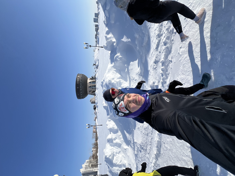 A group of four individuals is depicted engaging in outdoor activity on a bright, sunny winter day in Kazan, Russia. The scene is characterized by extensive snow cover forming drifts and mounds across the landscape. In the mid-ground, the distinct cauldron-shaped Kazan Family Center (Palace of Marriage) is prominently visible, establishing the location on the banks of the Kazanka River.
The person in the immediate foreground, positioned left of center, is taking a selfie. This individual is a male, wearing a black long-sleeved athletic top with a "FRAY" logo, a purple and white patterned balaclava or buff, and a black beanie, looking directly at the camera with a slight mustache. To his immediate right, another male is visible, wearing a blue jacket, black beanie, and red reflective sports sunglasses, appearing to move alongside the selfie-taker. To the far left, a person partially obscured, wearing a yellow and black top, is also in motion. To the far right, a fourth individual is partially visible from the waist down, wearing black athletic pants and light-colored running shoes, engaged in a running stride.
The ground is uniformly covered in thick, undisturbed snow, with a cleared or packed path where the individuals are situated. Several double-headed streetlights are scattered in the mid-ground. Tall residential or commercial buildings are visible in the distant background on both sides of the Kazan Family Center. The sky is clear and brilliant blue, casting sharp, elongated shadows on the snow from the people and objects, indicative of bright sunlight and a low sun angle.