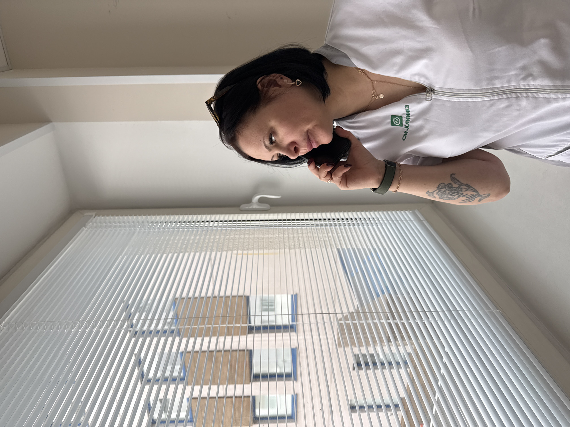 A woman in a white medical coat stands near a window with closed blinds. She is holding a black smartphone to her ear, seemingly engaged in a phone conversation. The logo "CM-Clinica" is visible on her coat. The window offers a partial view of a building across the street. The scene is likely set indoors, possibly in a clinic or office. The location is identified by the logo on the medical coat, which indicates the CM-Clinica chain in Moscow, Russia.