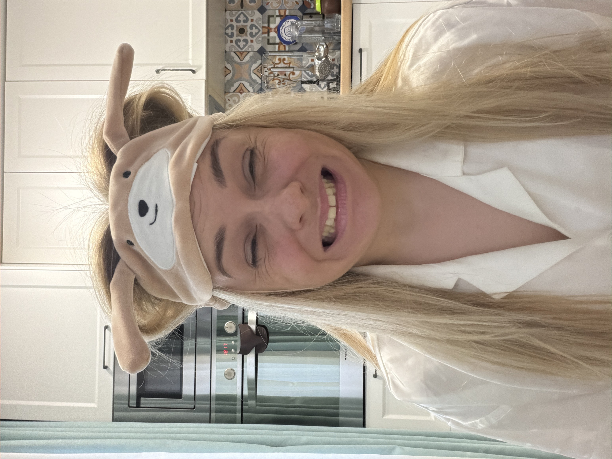 A woman with long blonde hair is depicted indoors, likely in a kitchen in Votkinsk, Russia. She is smiling broadly with her eyes squinted, suggesting happiness or laughter. She wears a light-colored sleep mask with animal-like ears (resembling a dog or bear) on her forehead, above her eyes, and a white, button-up shirt or pajama top.
The background reveals a kitchen with white upper and lower cabinets. A patterned tile backsplash, featuring blue, orange, brown, and white geometric designs, is visible between the cabinets. A stainless steel microwave or oven is integrated below the upper cabinets, with its digital display showing "8:17". Some kitchen items, including a metal rack with utensils or bottles, are hanging on the backsplash. A light blue surface, possibly a countertop or table, is in the foreground.
The overall scene conveys a playful and joyful moment, possibly in the morning given the sleep mask and the time displayed.