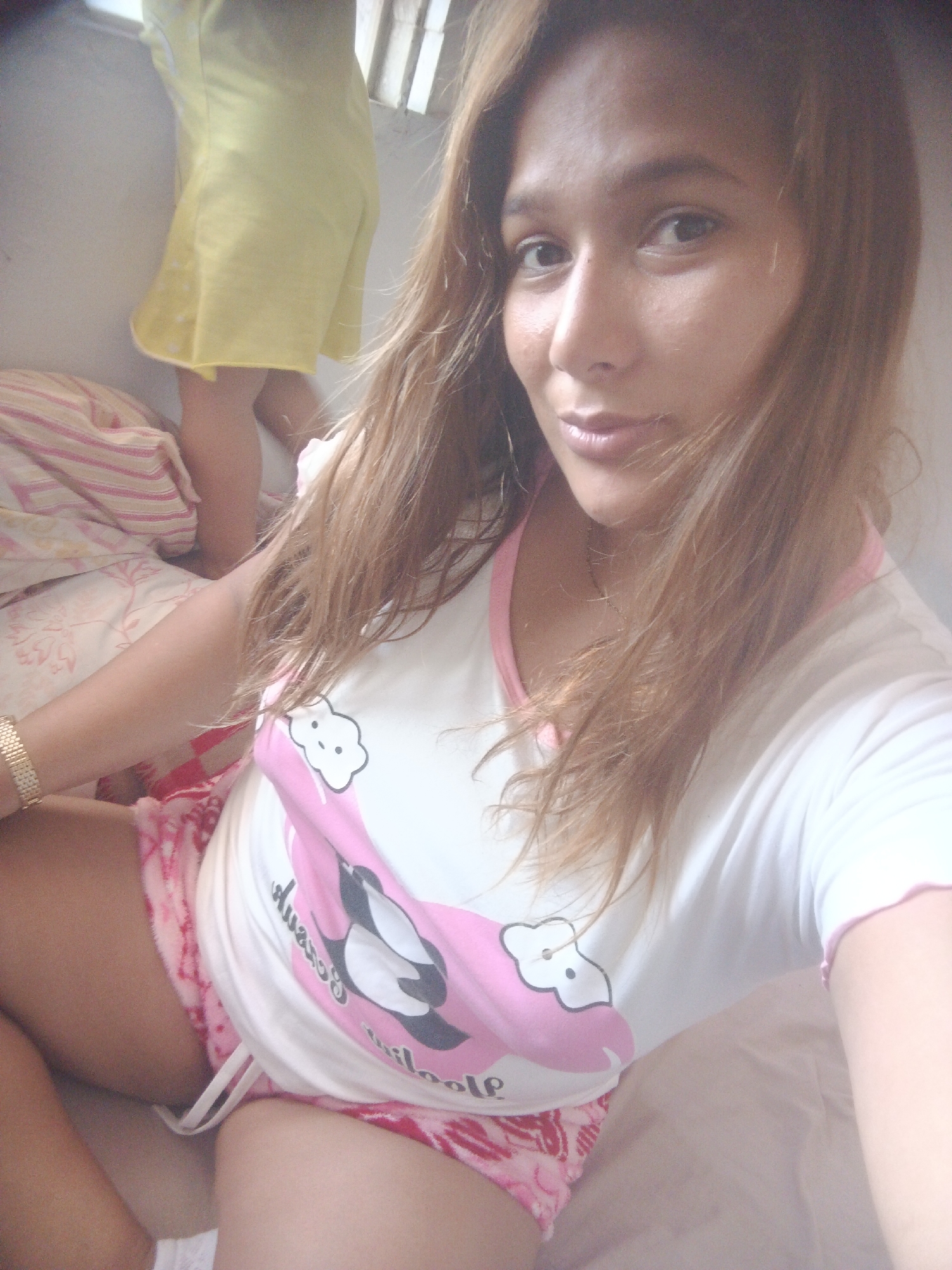 A person with long brown hair is seated on a bed with pink and white patterned sheets. The person is wearing a white t-shirt with a pink neckline and a pink design featuring a cartoon penguin and clouds. They are also wearing pink and white patterned shorts. A gold-colored bracelet is visible on their left wrist. In the background, another individual, possibly a child, is standing on the bed wearing a yellow dress. Light is entering from a window with white blinds. The scene appears to be indoors, likely in a bedroom. There are no details indicating the city of Caracas, Venezuela.
