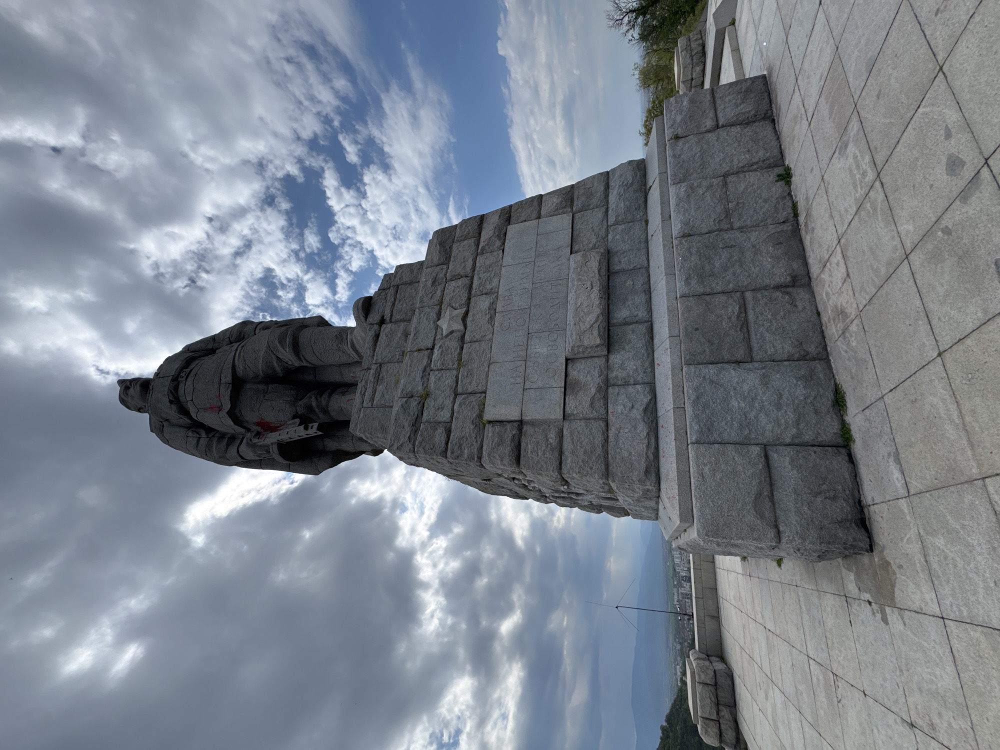 The image, taken in Plovdiv, Bulgaria, captures the imposing Alyosha Monument under a partially cloudy sky. The main subject is a towering stone statue of a soldier, depicted in a heroic, realist style, standing atop a massive stone pedestal. The statue itself shows traces of red graffiti on its midsection.
The monument's base is constructed from large, gray, hewn stone blocks. On one visible face of this pedestal, there is a carved inscription in Cyrillic, below which a five-pointed star emblem is also carved. The discernible text reads: "СЛАВА НА СЪВЕТСКАТА АРМИЯ ОСВОБОДИТЕЛКА," translating to "GLORY TO THE SOVIET ARMY, LIBERATOR."
The monument is situated on an elevated, paved platform made of lighter grey square tiles. Sparse greenery, likely weeds, can be seen growing at the base of the stone structures. The sky features a mix of bright blue patches and dynamic white and grey clouds. In the distance, a sprawling landscape of rolling hills or mountains and a cityscape are visible, suggesting the monument's commanding position overlooking the area. The scene is set during the daytime, with ample natural light. The photograph is taken from a low angle, emphasizing the monument's height and grandeur.