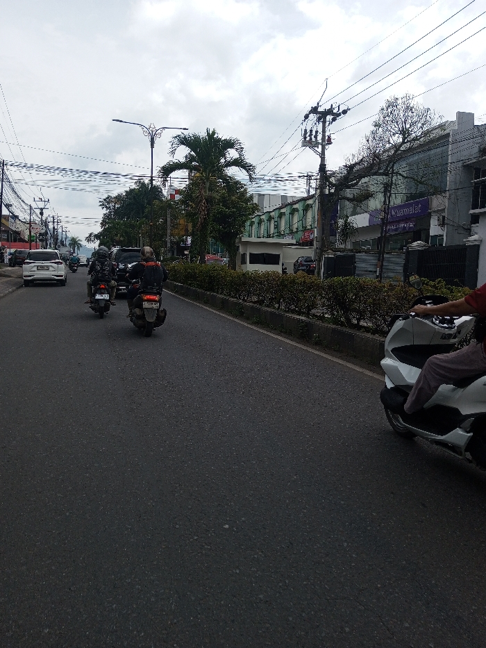 This image depicts a street scene under an overcast sky in Khajuripada, India. The asphalt road, appearing dry, features two traffic lanes moving away from the viewer, separated from the opposite direction by a raised concrete median strip topped with dark green hedges.
In the immediate foreground on the right, the front portion of a white scooter, likely a Honda PCX, is visible, operated by an individual wearing a red shirt and light-colored trousers. Ahead on the left lane, two dark motorcycles, both scooters, are in motion, heading away from the viewer. The lead motorcycle's driver wears a dark jacket with orange accents and a helmet, with license plate "B 5275 VBJ" visible. The second motorcycle's driver also wears a dark jacket and helmet, with license plate "B 6005 VME". Further back in the same lane, a white SUV is visible, followed by other vehicles.
The left side of the street is lined with commercial buildings and utility poles carrying numerous overhead power lines. A tall, ornate street light pole stands above the road in the mid-distance. The right side of the street features more buildings behind the median, including a prominent multi-story grey building with purple signage, bearing the text "Muamalat." Adjacent to it is a building with green paneling on its upper floors. A large palm tree and other assorted trees are visible throughout the scene, interspersed with buildings and utility infrastructure.