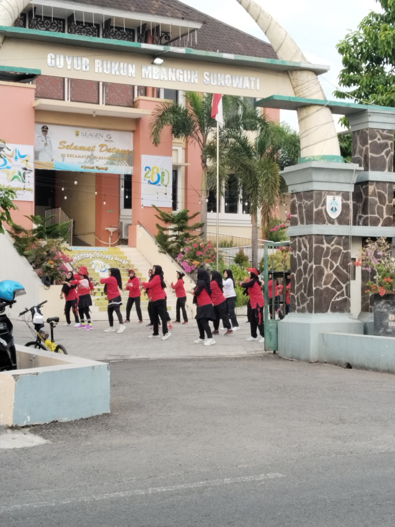 The image shows a group of women exercising outdoors in front of a building. The building has an arched entrance with the words "GUYUB RUKUN MBANGUN SUKOWATI" inscribed on it. There are banners and flags displayed on the building. The women are wearing red tops and black pants or skirts, and some are wearing headscarves. They appear to be performing some kind of aerobic exercise. There are trees and flowers around the building, and a street in the foreground. A blue helmet and a yellow bicycle are visible on the left side of the image. The scene suggests an organized group activity taking place in a public space.