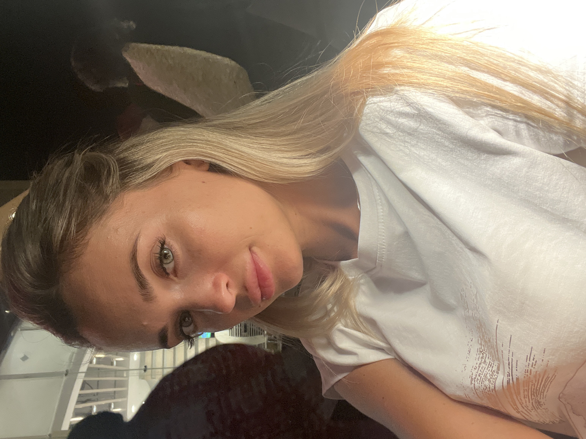 A blonde woman is captured in a close-up selfie, her gaze directed towards the camera. She is wearing a plain white t-shirt, which appears to have a subtle graphic design on the front. Her blonde hair is long and falls over her shoulders. The background is dark and out of focus, but hints of an indoor setting are visible, possibly a restaurant or cafe with some ambient lighting. There are blurred figures in the background to the left and right of the woman. The lighting suggests it is nighttime or an indoor location with dim lighting. The image appears to be taken in Sharm El Sheikh, Egypt, based on the provided context.