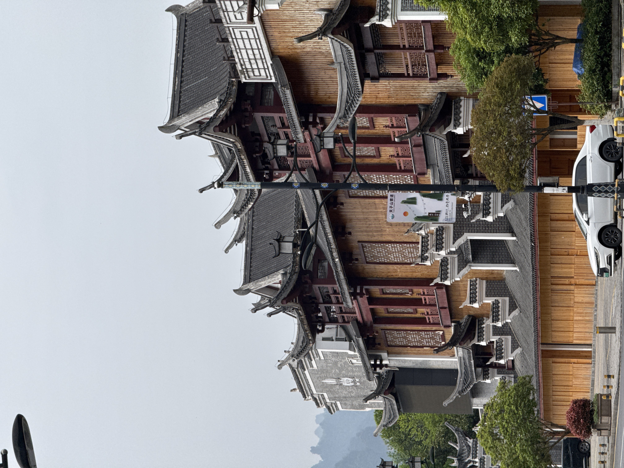 The image displays a street scene in Wulingyuan, China, characterized by traditional Chinese architecture under an overcast sky. A row of multi-story buildings, adorned with dark, ornate tiled roofs and upturned eaves, occupies the right side of the frame. These structures feature light brown wooden facades, reddish-brown pillars, and intricate lattice work on windows and balconies, with some sections incorporating grey brickwork.
Along the street in front of the buildings, a white car is parked. Several green trees and bushes are planted along the thoroughfare, enhancing the streetscape. A lamppost stands prominently, featuring decorative blue and yellow square tiles and a white banner. Further down the street, a blue rectangular road sign with white arrows is partially visible. In the background on the left, distant, hazy mountains rise against the pale, diffused light of the sky.
The scene appears to be static, capturing the architectural beauty and setting during daytime with soft, uniform lighting, indicating an overcast or cloudy weather condition. There are no people visible, and the activity is limited to parked vehicles, suggesting an everyday street environment or a tourist area.
Visible text on the banner attached to the lamppost includes "天子山索道" (Tianzi Mountain Cableway) in Chinese, immediately followed by its English translation, "TIANZI MOUNTAIN CABLEWAY". Smaller Chinese characters are also visible below, likely indicating "张家界市" (Zhangjiajie City) and "核心景区" (Core Scenic Area), pinpointing the location within the broader Zhangjiajie region.