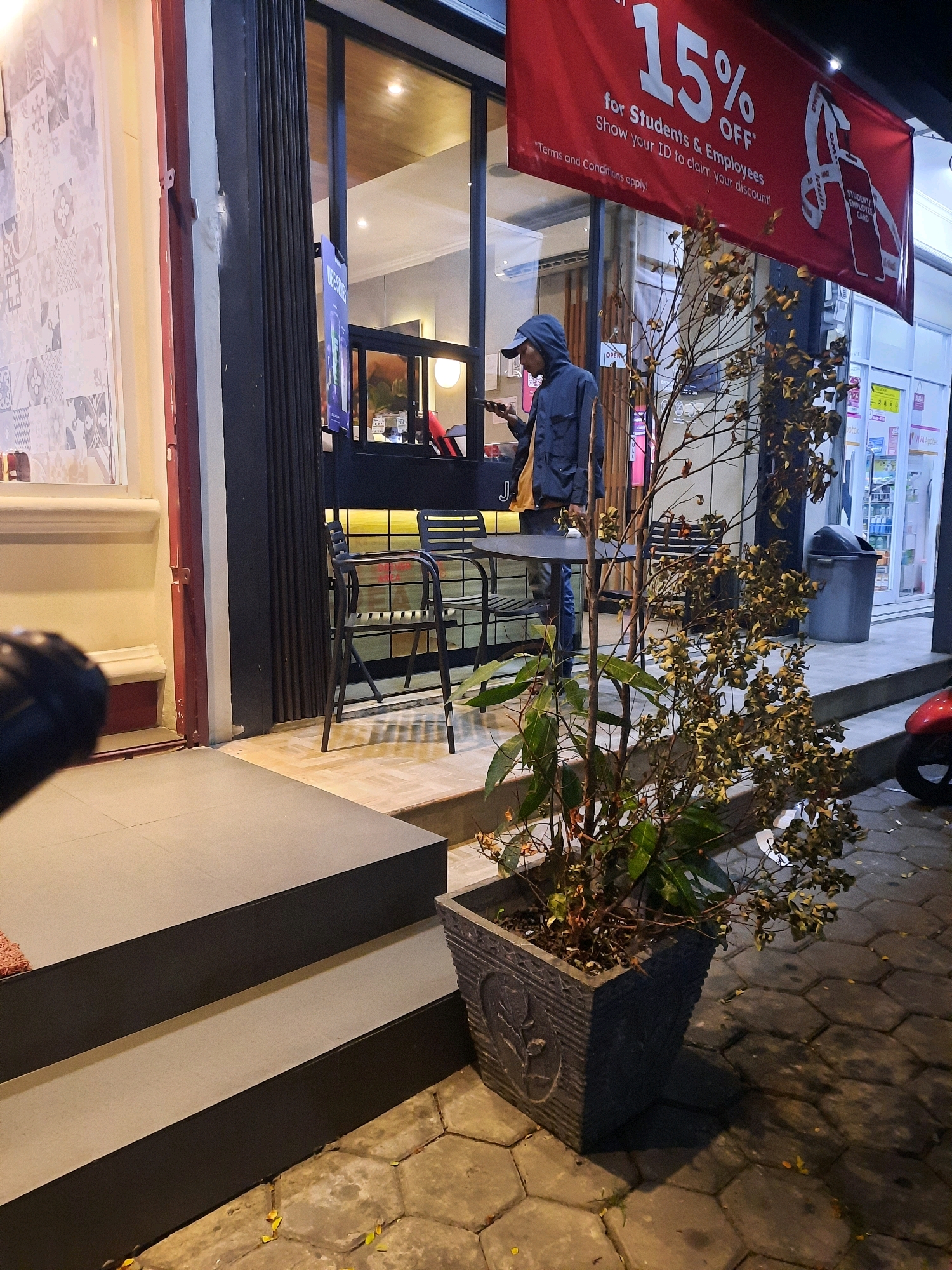 The image depicts an outdoor scene at night in front of a building in East Jakarta, Indonesia. A person wearing a hooded jacket and a cap is standing outside, looking at their phone. In the background, there's a red banner advertising "15% OFF for Students & Employees". The building appears to be a shop or restaurant with large glass windows and a dark-colored retractable gate. Outside, there are tables and chairs, a large potted plant with dried foliage, and a grey trash bin. The ground is paved with interlocking hexagonal stones, and there are some fallen yellow leaves. To the left of the scene, there are dark gray steps leading up to the building's entrance. A portion of a red motorcycle is visible on the far right. The lighting suggests it is evening or night.