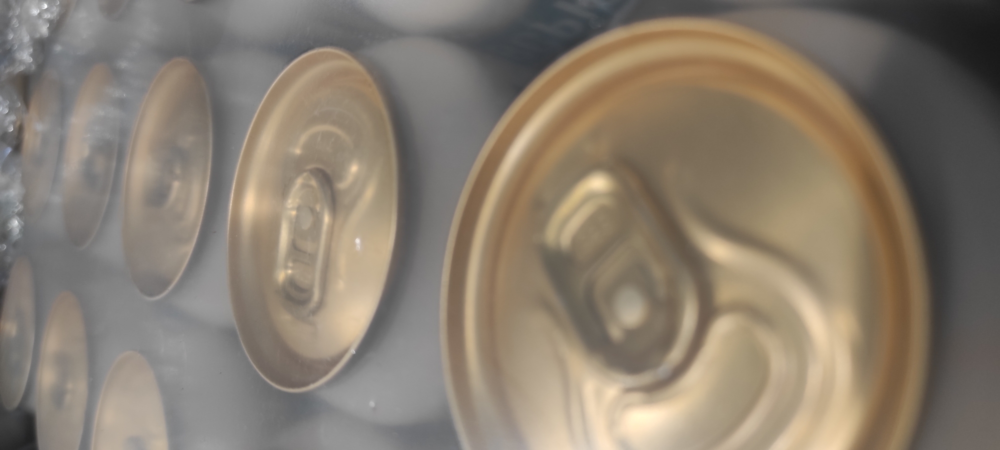 The image shows a close-up, top-down view of several gold-colored aluminum cans arranged in rows, likely inside a refrigerator or cooler. The cans have a metallic sheen and a slightly matte finish, with visible pull-tabs on their tops. The perspective is shallow, with the foreground can sharply in focus and the background cans progressively blurred. The overall color palette is dominated by shades of gray, silver, and gold, with a soft, diffused light. There is no visible indication of the city of Kamianske, Ukraine, or any elements that would suggest a specific location. The image focuses solely on the arrangement and appearance of the beverage cans.