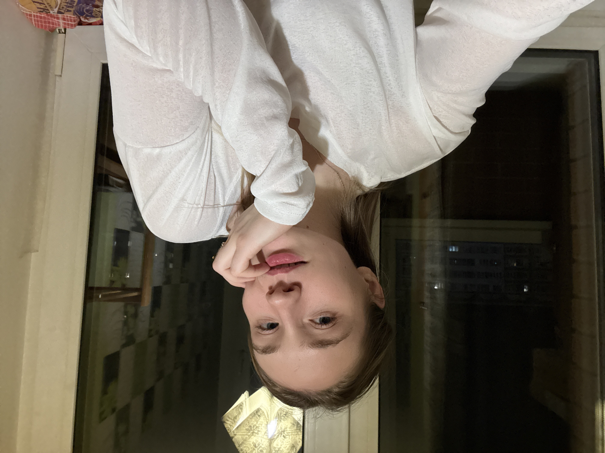 A young woman with fair skin and light brown hair is posing for a selfie in front of a window. She is wearing a white, long-sleeved shirt and a delicate necklace. Her right hand is touching her chin. The window reflects a cityscape, likely Izhevsk, Russia, due to prior knowledge. The light inside and reflected outside suggests nighttime.