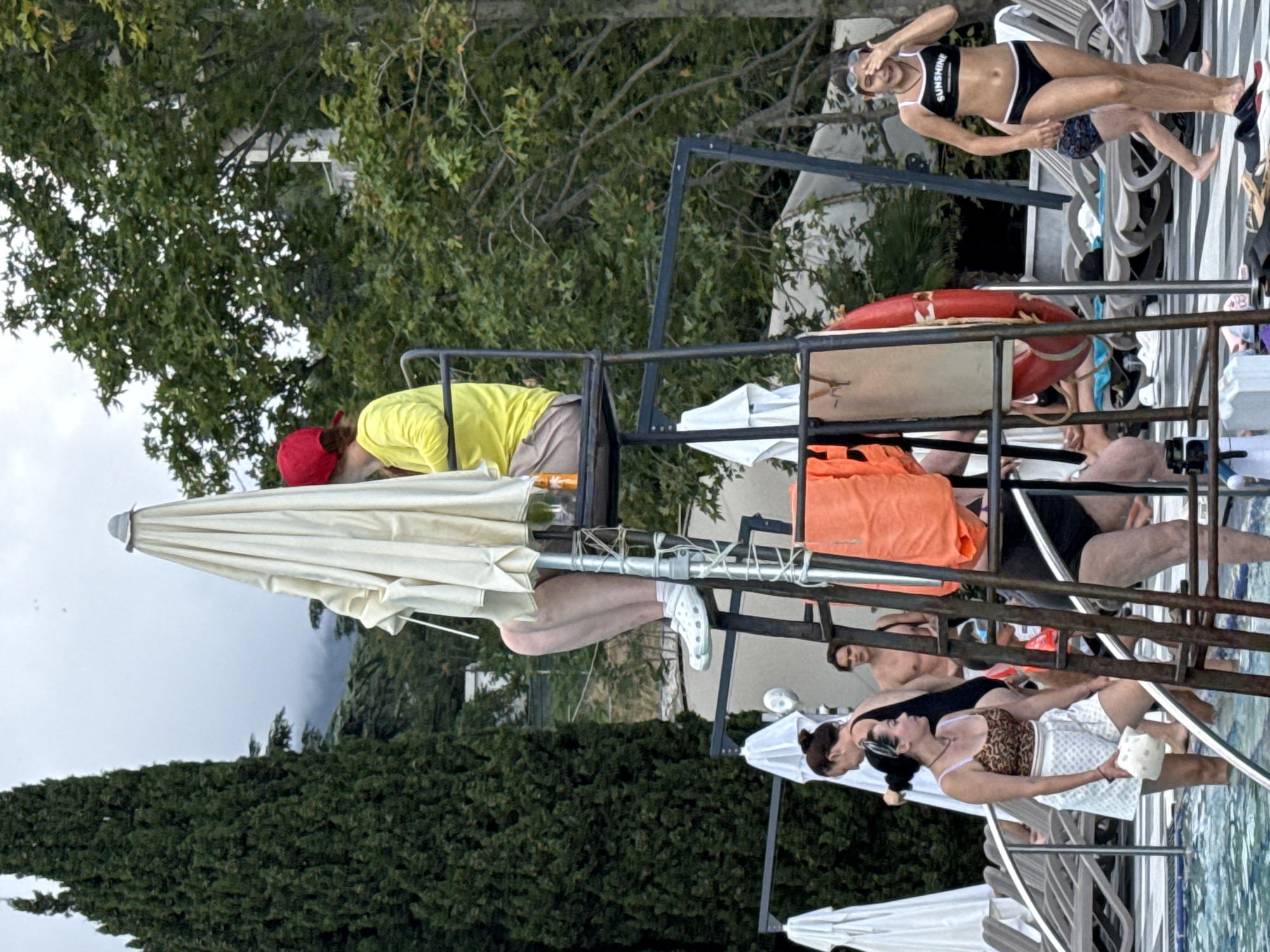 The image depicts an outdoor swimming pool area. In the foreground, several people are visible in and around the pool. A lifeguard, wearing a red cap and yellow long-sleeved shirt, is seated on a high chair under a large white umbrella. Other individuals in swimwear are standing or wading in the pool. To the right, a young girl in a black bikini with white trim, wearing goggles, is standing near the edge of the pool, appearing to wipe her eyes. Loungers and beach umbrellas are set up around the pool area. Lush green trees and foliage form the backdrop, with a hint of mountainous terrain and overcast sky visible in the distance. The setting suggests a resort or hotel pool, likely in a warm climate, given the presence of swimwear and the overall ambiance. The specific location in Массандровский поселковый совет, Ukraine, is not explicitly identifiable from visual cues alone, but the natural environment with its trees and potentially hilly terrain could be consistent with the region.