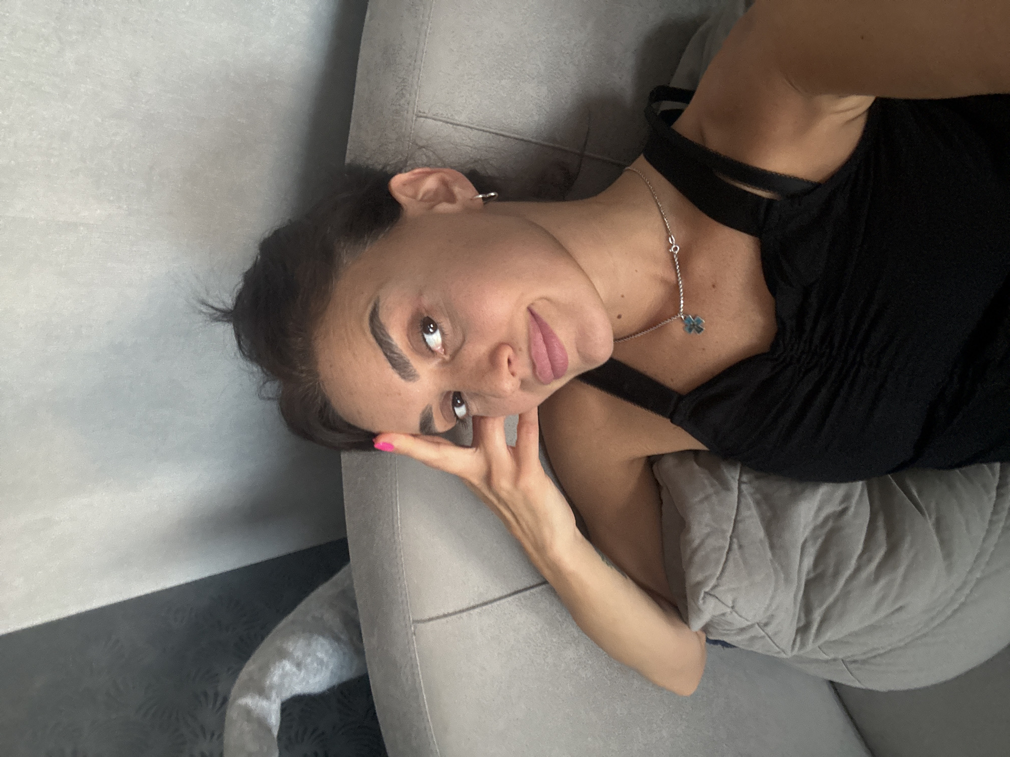 An adult female is lying down on a sofa and resting her head on her hand. She is wearing a black tank top and a silver necklace with a blue pendant. The background appears to be a wall with a textured pattern. The image was taken in Tbilisi, Georgia.