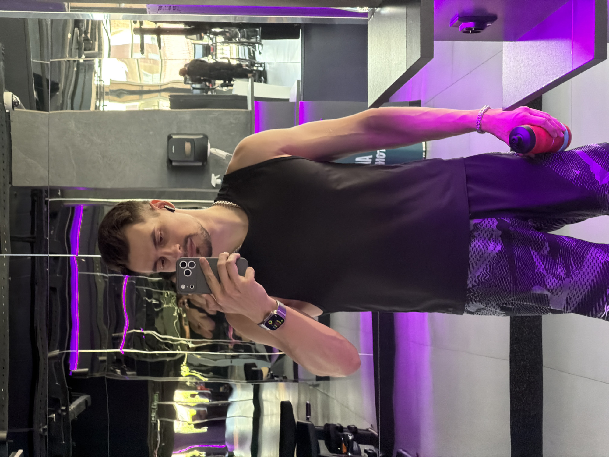 This media file shows a young man taking a selfie in a gym. He is wearing a black tank top and patterned shorts. He is holding a red water bottle in his right hand and his phone in his left. He appears to be wearing a watch on his left wrist. The gym has a modern aesthetic with large mirrors and purple lighting. The reflection in the mirrors shows gym equipment. The image is taken in portrait mode and is likely indoors. There is no information about the time of day or weather. The man's expression is neutral, and there are no strong emotional cues. There is no visible text in the image.