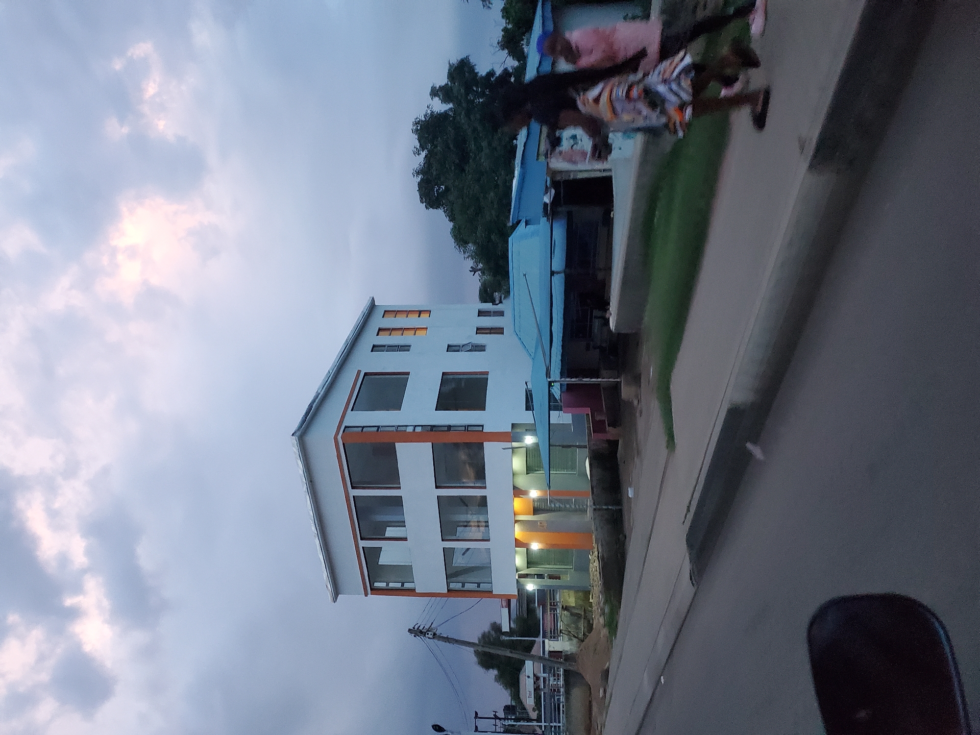 The image depicts a street scene in Gura Top, Nigeria, at dusk. A modern, three-story building with white and orange accents and large glass windows dominates the right side of the frame. In front of the building, a blue-tarped structure and a small open-air stall are visible. Two individuals, one wearing a patterned dress and the other a pink top, are walking on the sidewalk. The street is paved, with a concrete curb separating it from the sidewalk. Trees and power lines are present in the background. The sky is overcast with hints of pink and yellow light.