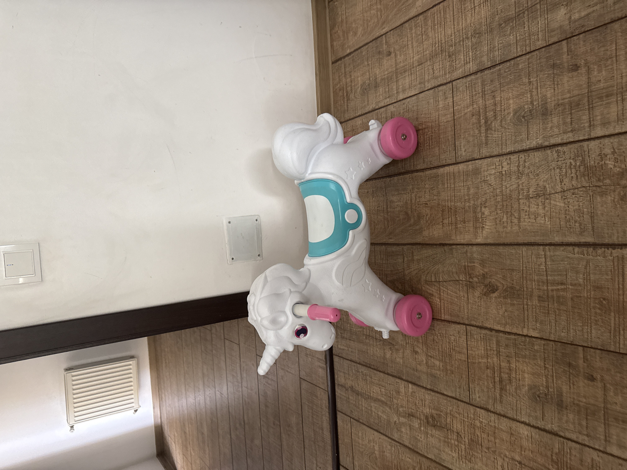 A white and turquoise toy unicorn with pink wheels and pink handlebars is positioned on a wooden floor. The unicorn is facing left and has a horn, eyes, mane, and tail. A white wall is behind the toy, and a doorway with a dark brown frame is to the left, revealing a glimpse of a white radiator in another room. The floor is made of brown wooden planks. The image appears to have been taken indoors, possibly in a residential setting in Chișinău, Moldova. There are no people present in the image.