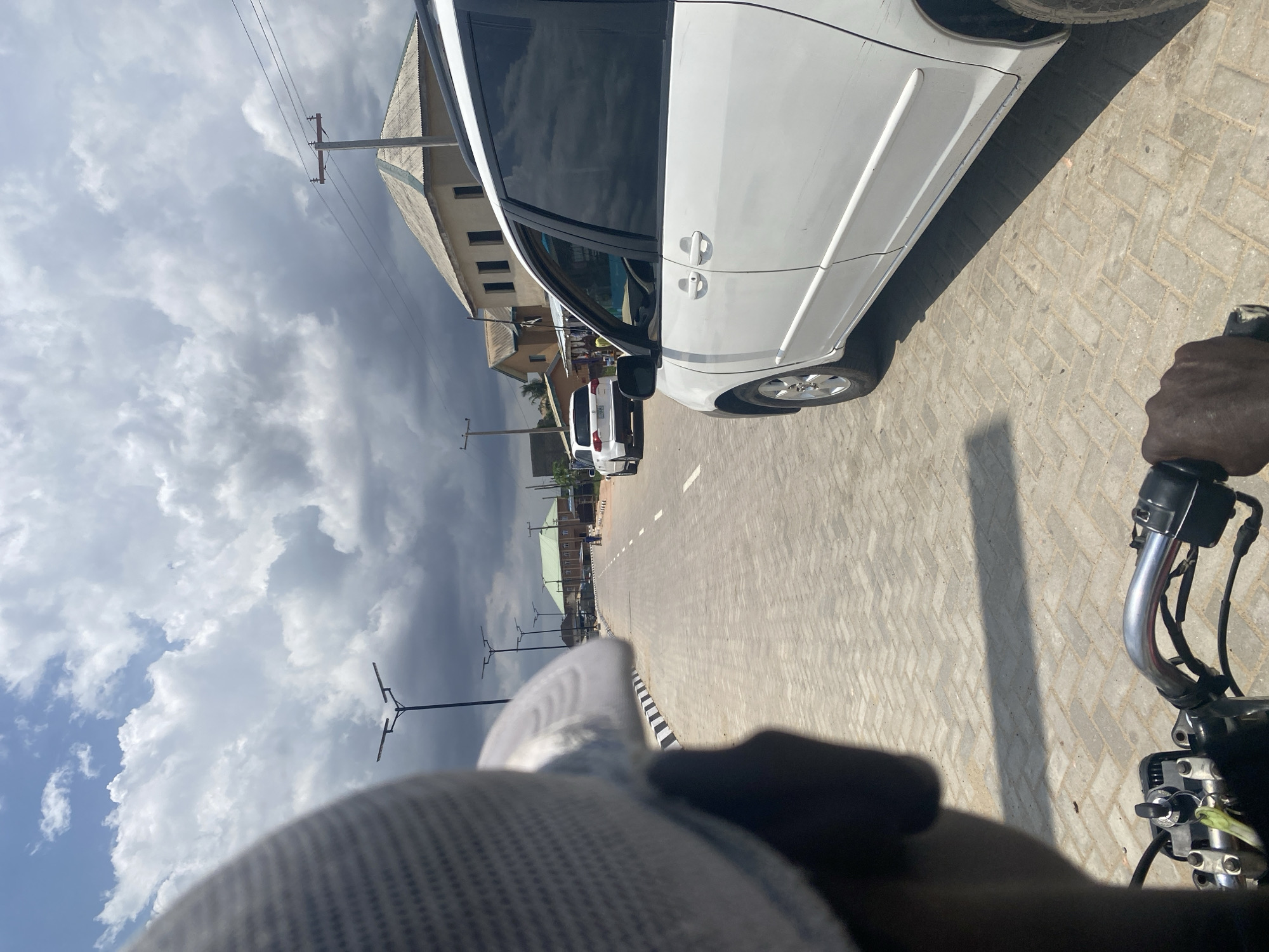 The image depicts a street scene from a low, first-person perspective, possibly from a person riding or stopped on a motorcycle or scooter. In the bottom right foreground, a dark-skinned hand is visible gripping the handlebar of a vehicle, showing part of the throttle and control mechanisms. The metallic handlebar reflects light. In the bottom left, the brim and mesh texture of a cap or hat are visible, suggesting the perspective of the wearer.
Immediately to the left of the handlebar and hand, a light-colored paved road made of interlocking pavers stretches into the distance. A white dividing line runs along the center of the visible road segment. A curb with black and white stripes separates the road from what appears to be a sidewalk or shoulder. Several modern street lights with dark, T-shaped tops are spaced out along the road.
A white passenger vehicle, possibly a minivan or SUV, is parked or moving slowly on the left side of the road, partially obscuring a building behind it. The car has dark tinted windows. Further down the road, another white car is visible. In the background on the left, several buildings with light-colored walls and dark roofs are present, along with utility poles and power lines strung across the sky.
The sky is largely overcast with dramatic, heavy grey and white clouds that appear denser on the left side, hinting at a potentially stormy or heavily clouded day. A patch of clearer blue sky with lighter clouds is visible in the upper left corner. The lighting casts some shadows, suggesting it's daytime, possibly late morning or afternoon. No specific activity beyond being on a street is evident, and there is no visible text.