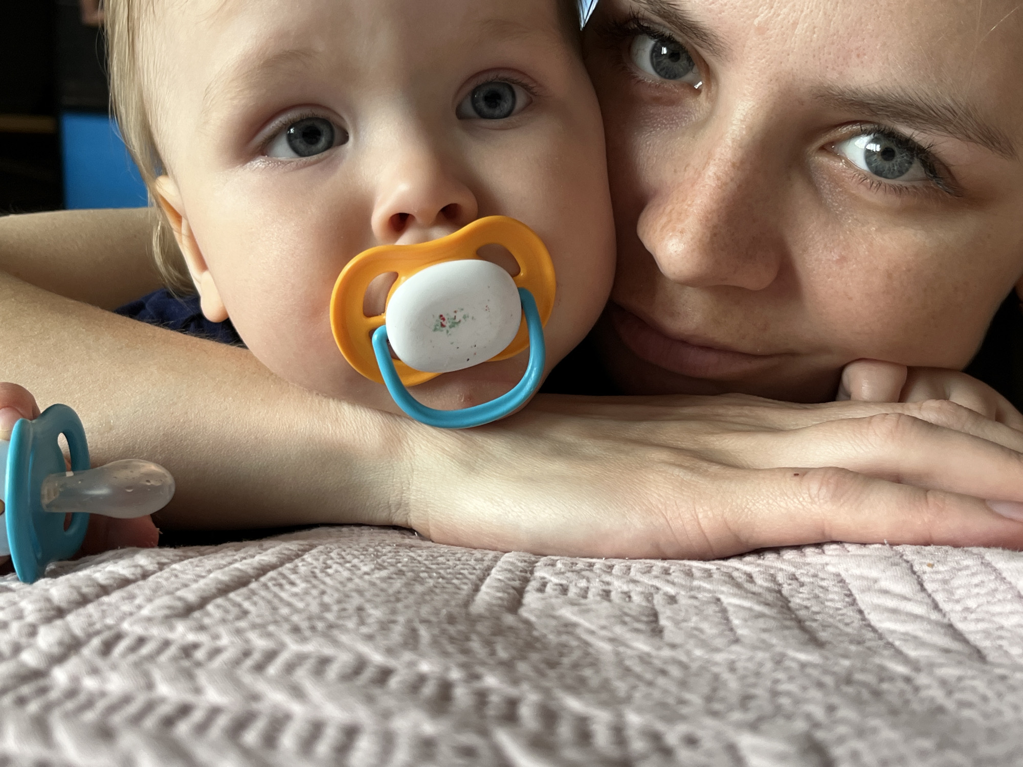 A close-up shot captures a baby with a pacifier nestled against an adult woman. The baby, positioned to the left, has fair skin, light hair, and striking blue eyes. The pacifier is predominantly white with a blue handle and an orange shield. The woman, on the right, has fair skin, freckles, and light-colored eyes. Her face is positioned near the baby's, suggesting a close relationship. Both are resting on a pink textured surface, possibly a blanket or bedding. A second pacifier, also blue, lies to the left of the frame. The background is blurred and indistinct. There is no information to support that this image was taken in Chaikovsky, Russia.