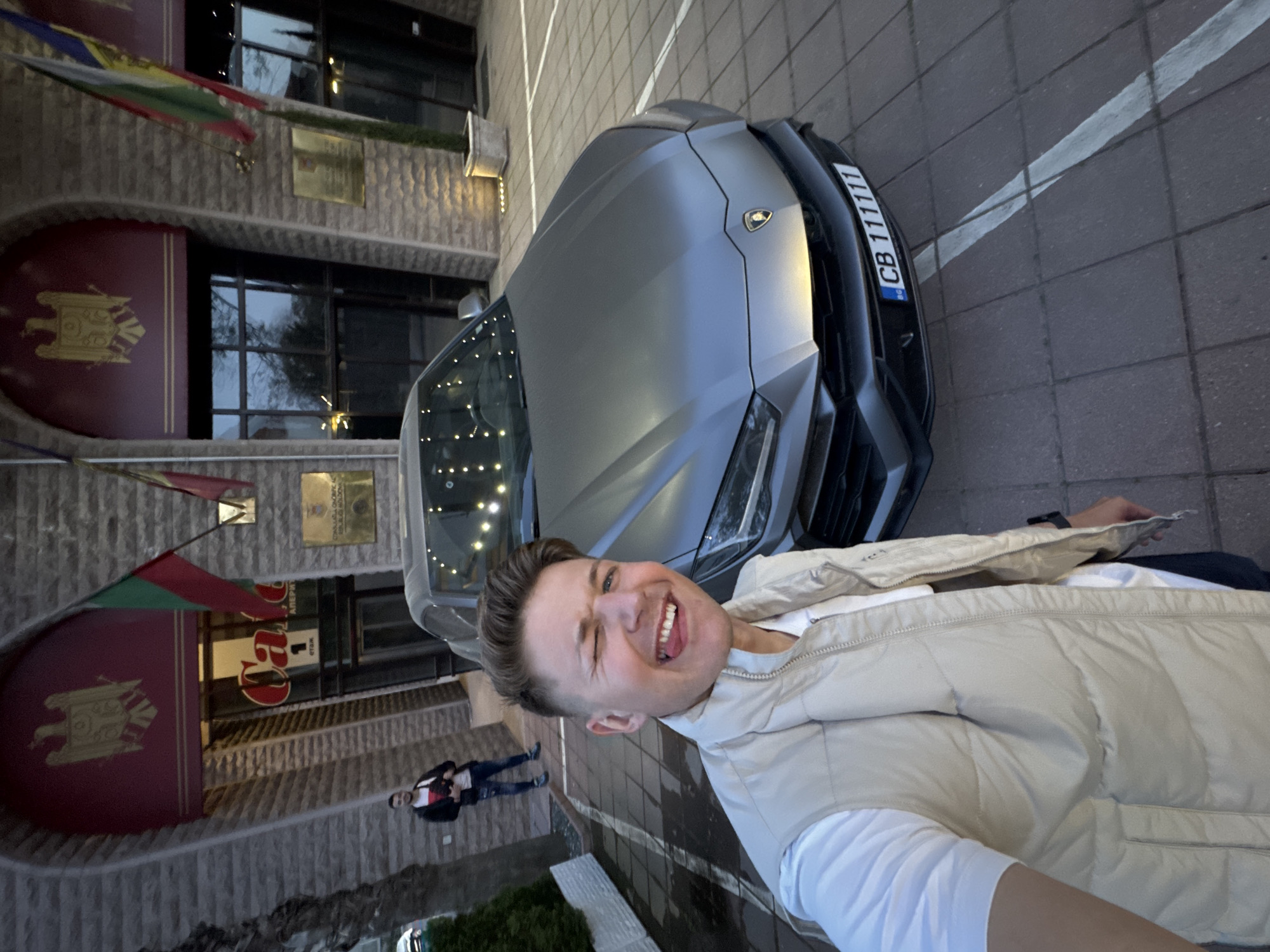 A young man takes a selfie in front of a grey Lamborghini Urus SUV, parked on a paved surface. He is smiling widely with a winking eye and his tongue slightly extended. He wears a white t-shirt and a light beige puffer vest. The Lamborghini emblem is visible on the car's hood. The car's license plate, "CB 111111," prominently displays the "BG" country code, indicating Bulgaria, with "CB" signifying Plovdiv.
Behind the man and the car is a multi-story building featuring a textured, stone-like facade and dark-framed windows. Above one of the entrances, a large maroon, curved awning or sign is adorned with the golden Coat of Arms of Moldova. Below this, a partially visible sign reads "Casa 1" and "ЕТАЖ" (floor). A golden plaque mounted above the awning, though its Cyrillic text is difficult to fully decipher, likely indicates "КОНСУЛСТВО МОЛДОВА" (Consulate of Moldova). Two flags are displayed on the building: one featuring red, yellow, and blue sections (consistent with the Moldovan flag) and another primarily green and red.
Another person, dressed in a dark jacket and jeans, is visible in the background, standing further back near the building's entrance. The scene appears to be set during late afternoon or early evening. The overall setting places the event in an urban environment, specifically Plovdiv, Bulgaria, as indicated by the license plate and the probable Moldovan consulate.