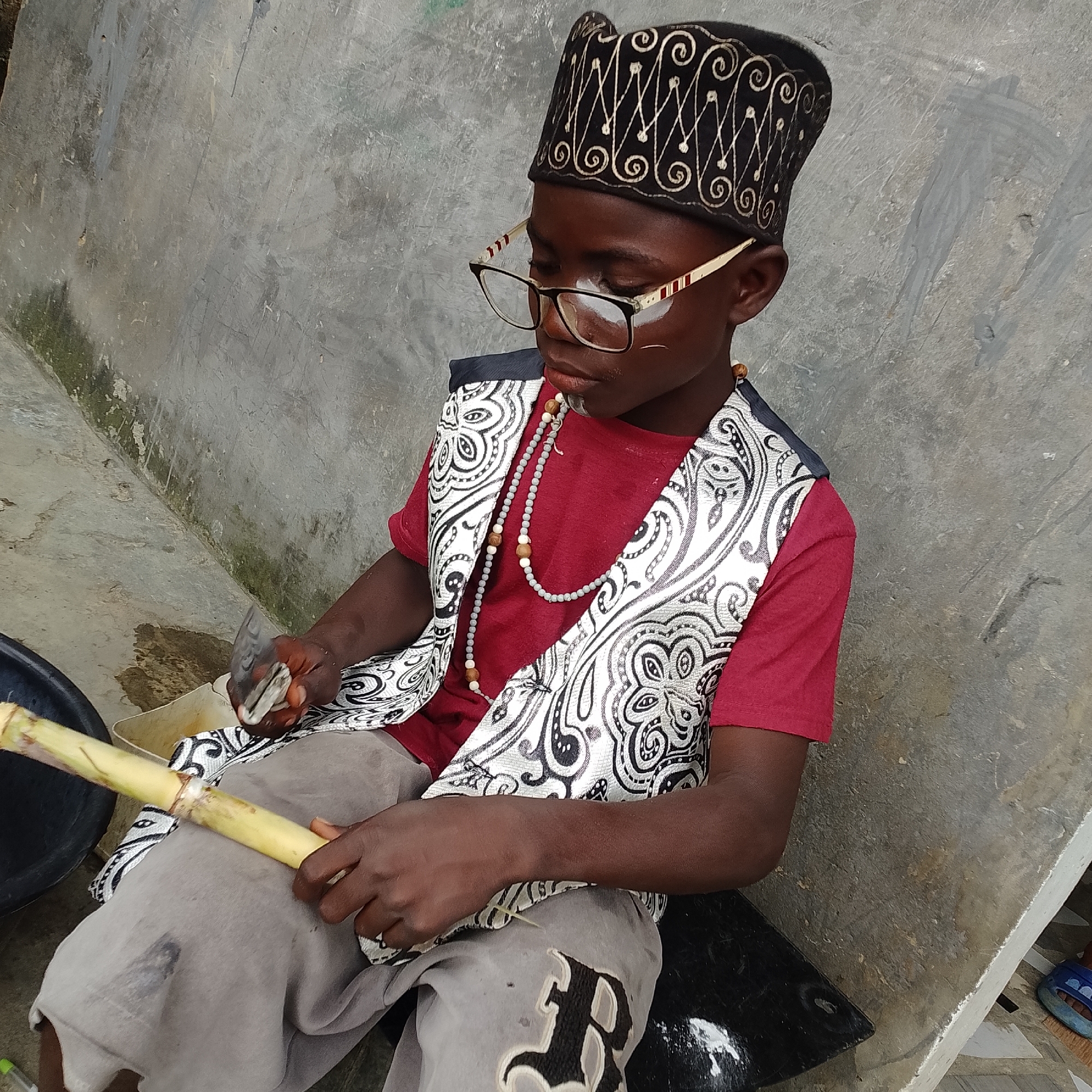 A young boy, appearing to be of Nigerian descent and dressed in a traditional cap and vest over a red shirt, sits on a black surface next to a concrete wall. He is wearing glasses and has white markings on his face. In his hands, he holds a piece of sugarcane and a knife, suggesting he is in the process of preparing it. A black bowl is visible to his left. The environment appears to be outdoors, with a rough, textured concrete wall forming the backdrop. The lighting indicates it is daytime. The scene is likely located in a residential or communal area in Ado, Nigeria.