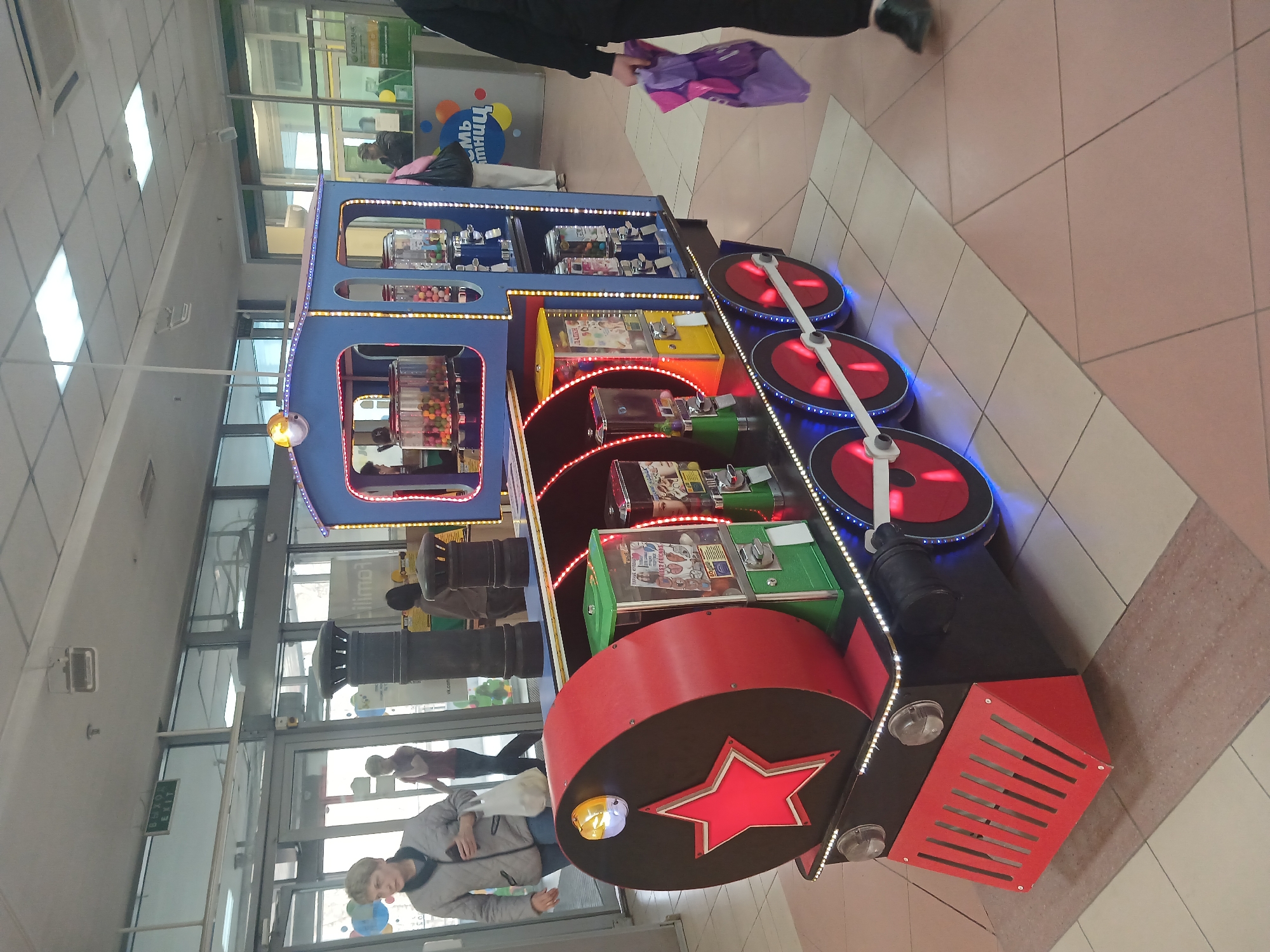 The image depicts a brightly colored, train-shaped vending machine in what appears to be a shopping mall or similar commercial space. The machine is blue, red, and black with LED lighting. It features several compartments filled with gumball machines, toys, and other vending items. A large red star is prominently displayed on the front of the "train." The floor is tiled in a geometric pattern with pink and white tiles. People are seen walking in the background near a glass entrance/exit of the building. Based on the Cyrillic text on the sign in the background ("Птицы"), this is in Russia. Judging from the Cyrillic text, it is likely located in the city of Perm.