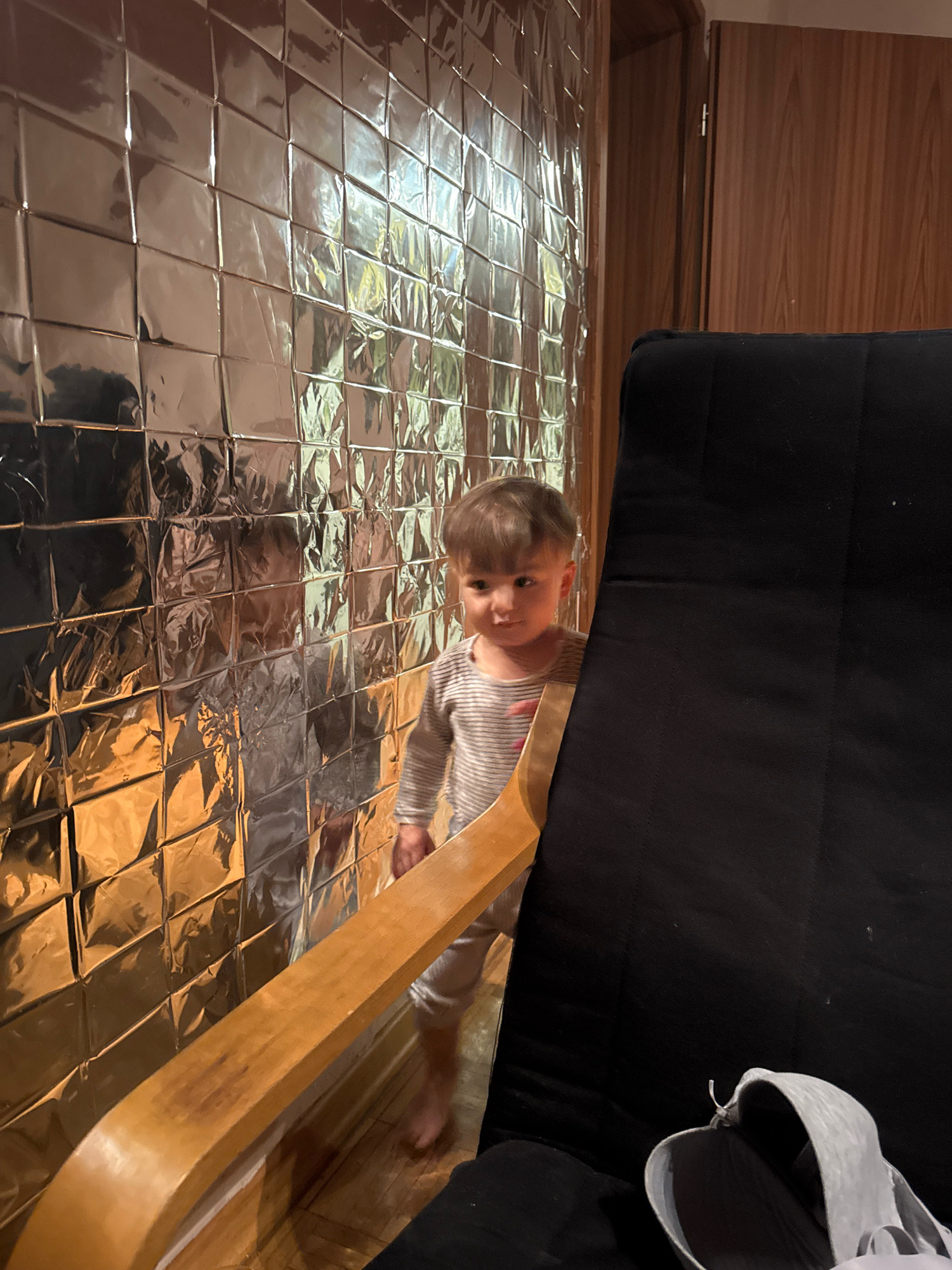 A young child, with light brown hair, is partially visible in the center of the frame, peeking from behind a large black chair. The child is wearing a horizontally striped long-sleeved shirt and light-colored pants. They are standing on a wooden floor, barefoot. To the left of the child, a wall covered in reflective, square tiles dominates the background, creating a shimmering, distorted effect. The tiles appear to be metallic and are arranged in a grid pattern. To the right of the child and chair, a wooden door frame and a portion of a wooden door are visible. The overall lighting is warm, with reflections from the tiled wall casting golden hues. This scene appears to be an interior shot, possibly in a domestic setting. The specific location in Überherrn, Germany, cannot be determined from the visual information alone.