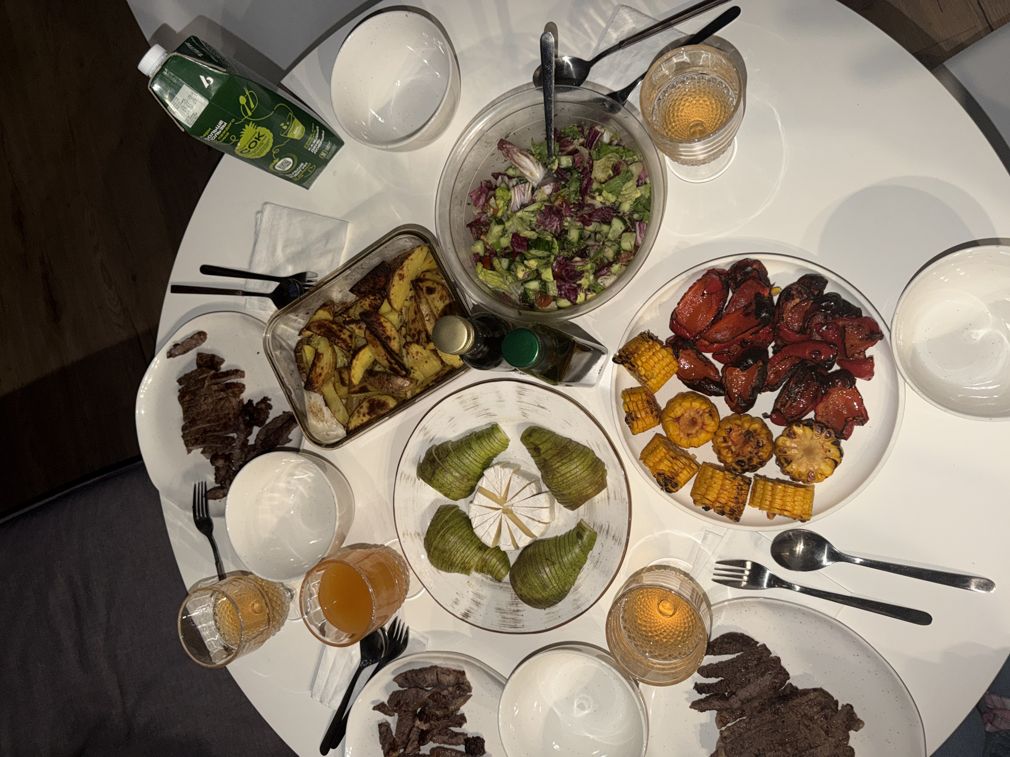 The image captures a top-down view of a round table laden with food. The scene presents a variety of dishes, including a salad in a clear bowl with utensils, sliced steak on two separate white plates, roasted potatoes in a glass dish, grilled corn and red peppers on a white plate, and a plate featuring brie cheese surrounded by sliced pears. There are also four glasses containing different colored liquids, a carton of what appears to be juice, and several empty white bowls scattered around the table. Forks and spoons are placed near each plate. The background suggests an indoor setting with a dark chair visible on the left side of the frame. The presence of a carton with Cyrillic text on it suggests that the location might be in Russia. Considering the image, it is impossible to conclusively determine the exact location is in the city of Krasnoyarsk.