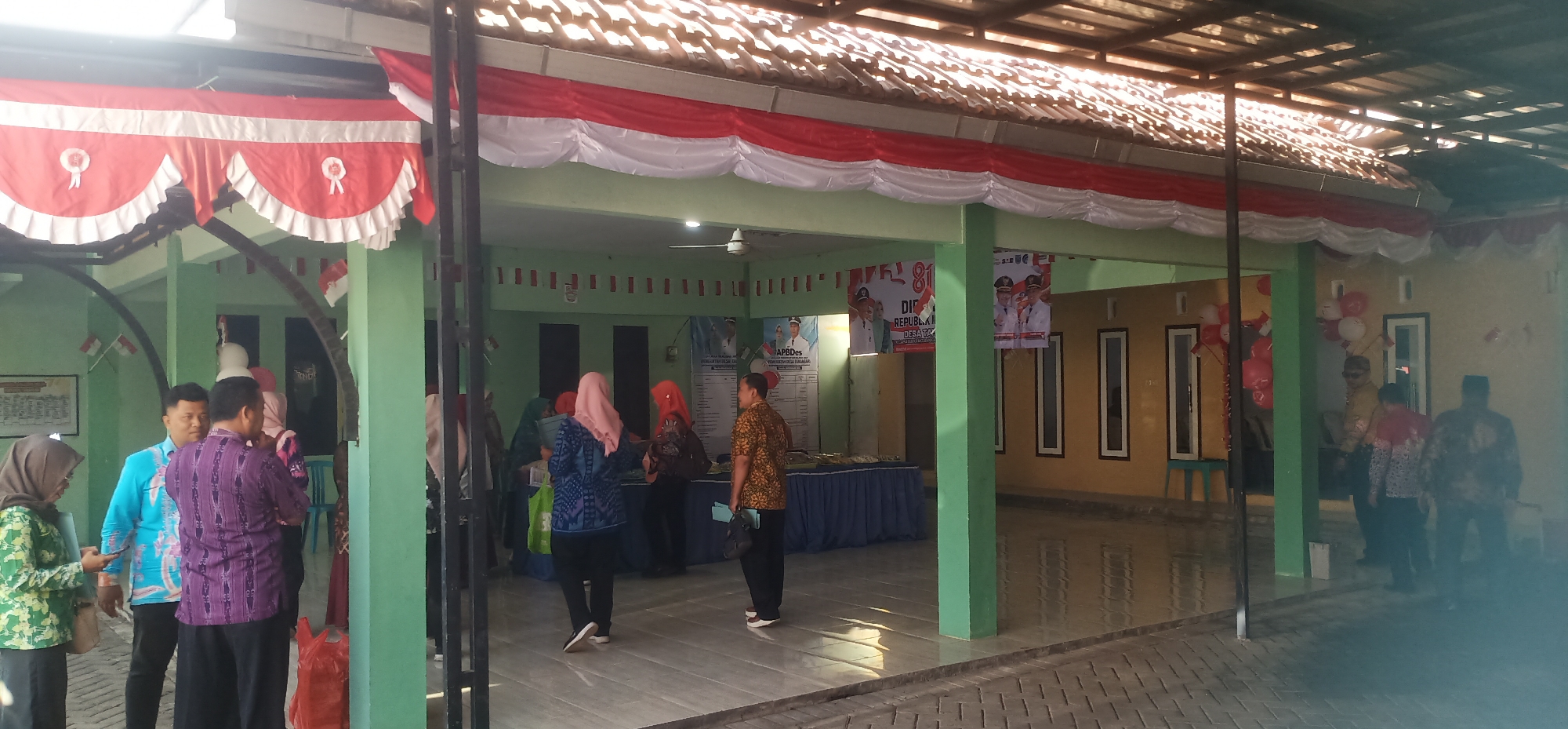 This image depicts a gathering at what appears to be a community building or event space in Tamansari, Indonesia, likely for a local government or festive occasion. The building has a light green exterior and a tiled roof with visible wooden beams. Red and white banners, possibly representing the Indonesian flag, are draped along the roofline and on poles. Several people are present, mostly dressed in casual attire with some individuals wearing batik shirts, a common Indonesian garment.
In the foreground, to the left, a woman in a green floral patterned outfit and headscarf is looking at her phone. Next to her, a man in a bright blue patterned shirt is talking. Further into the structure, several individuals are gathered, some standing in conversation and others appearing to be looking at displays or information boards. Two large posters featuring men in official attire, likely local leaders or government officials, are visible on the green wall, along with text that appears to be in Indonesian. Red and white balloons are tied to a structure on the right side of the image. The floor inside the building is reflective, suggesting it is paved or tiled and possibly wet, or polished. The overall impression is a community event with a festive and official atmosphere.