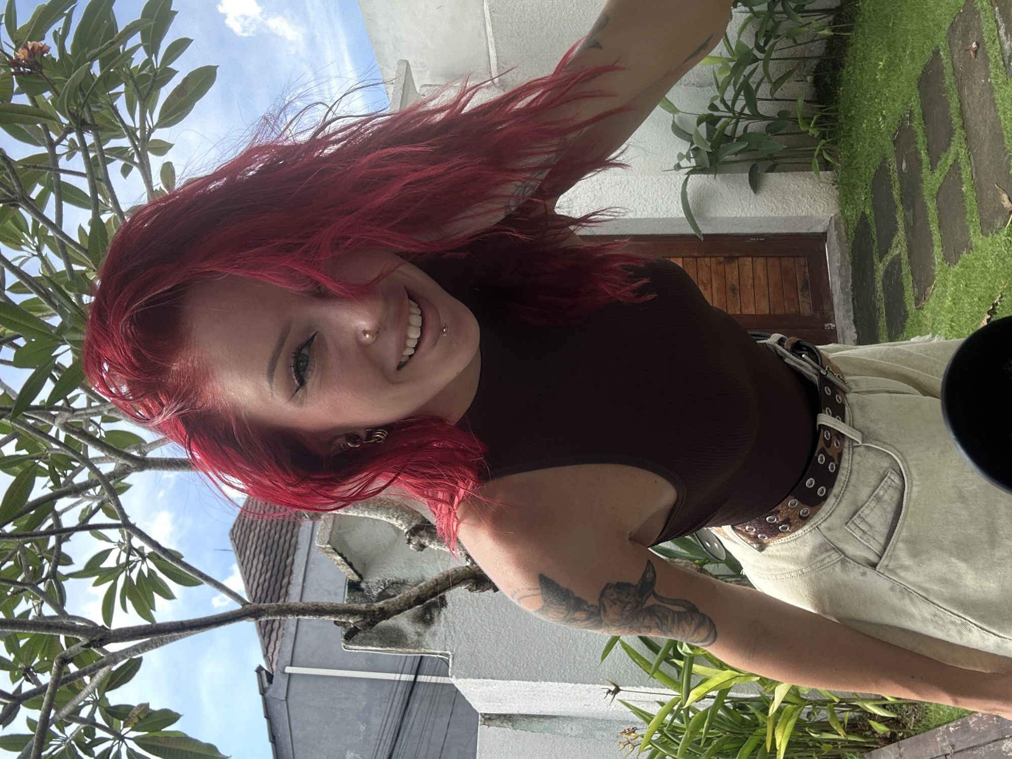 An outdoor portrait captured with a tilted perspective shows a young person with bright red, long hair flowing around their face. They are smiling broadly at the camera, revealing teeth, with a nose ring, lip piercing, and ear piercings visible. The individual wears a dark brown ribbed tank top and light-colored, high-waisted pants secured with a patterned belt featuring eyelets, possibly leopard print. Tattoos are visible on both arms, including what appears to be an animal design on the right arm.
The setting is a sunny outdoor space, consistent with a tropical location like North Kuta, Indonesia. Lush green foliage, including a tree with large, broad leaves (possibly a frangipani), frames the upper and left portions of the image. The background features a light-colored building with a wooden door and small green plants growing along its base. The ground shows a pathway of irregular stepping stones embedded in green grass. The sky is bright blue with scattered white clouds, indicating clear weather during the daytime, likely midday or afternoon. There is no visible text in the image.