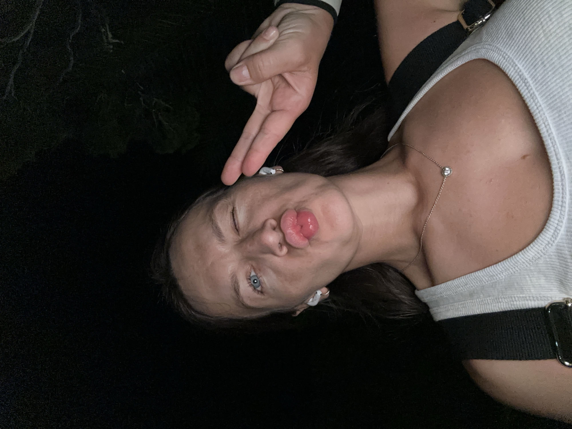 A woman with blue eyes and dark hair is taking a selfie at night. She is winking and puckering her lips, while holding up two fingers in a peace sign with her right hand. She is wearing a white tank top and a silver necklace with a small pendant. She has white earbuds in her ears. The background is dark and appears to be outdoors, possibly in a wooded area. The image was taken in Budva, Montenegro.
