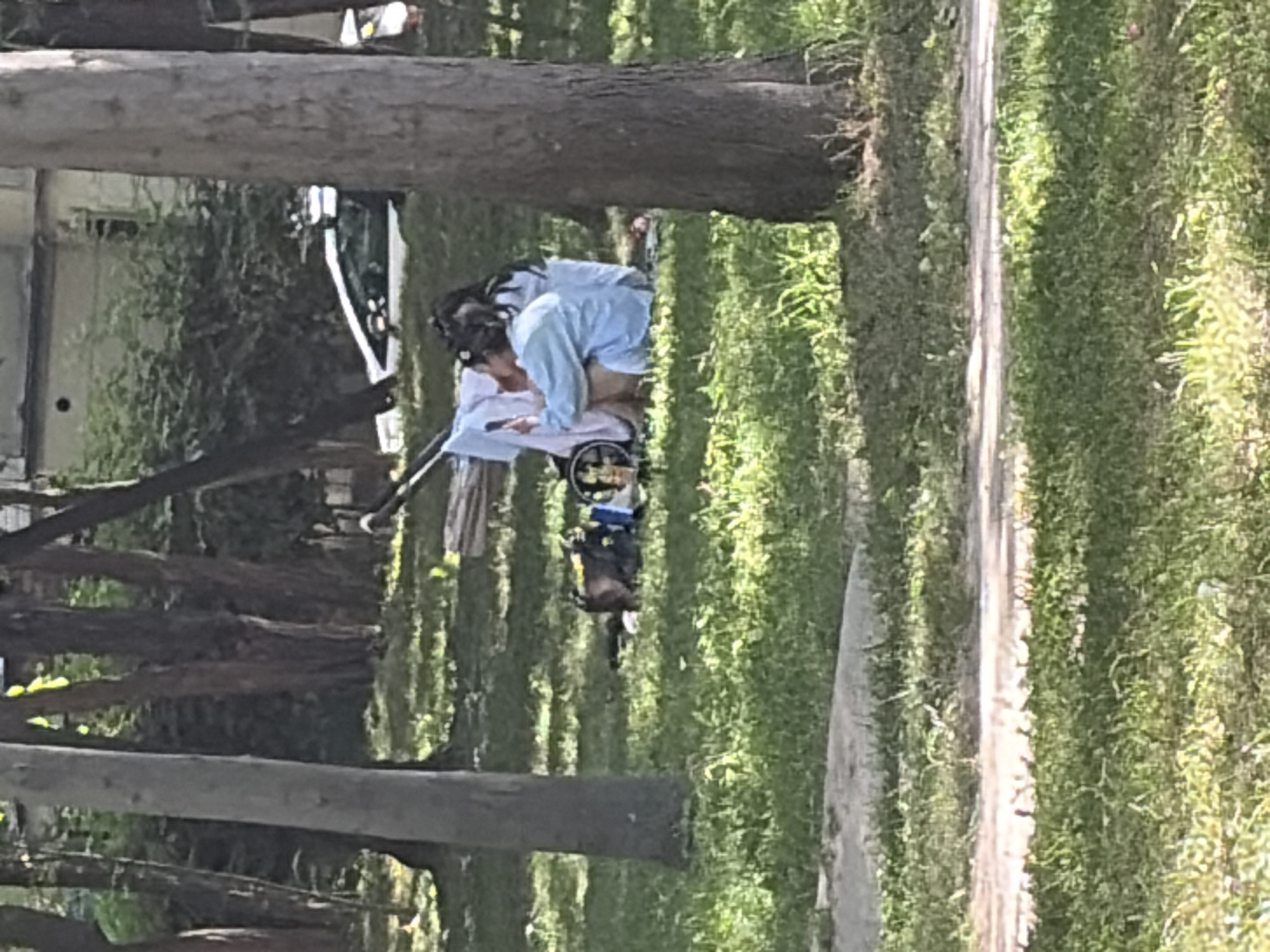 The image depicts a park scene with two individuals sitting on the grass. A stroller is visible behind them, with some clothing draped over it. One person, wearing a light blue shirt, is holding a smartphone and appears to be looking at it. The other person, also in a light blue garment, is seated next to them. The ground is covered in grass, and trees are present in the background, with a building and a white car partially visible further back. The setting appears to be a sunny day.