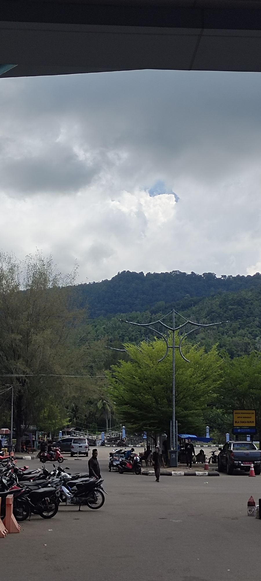 A vertical, eye-level, outdoor, full-shot photograph captures a cloudy day in Sabang, Indonesia. The scene is dominated by a parking lot filled with numerous motorcycles, some parked and others with people interacting around them. To the right, a black pickup truck with a license plate that reads "BL 9170 JX" is parked near an orange traffic cone. Further in the background, several cars and more motorcycles are visible, suggesting a bustling area. Beyond the parking lot, a lush green forest covers a hillside that slopes upwards to a mountain range under a heavy, overcast sky with patches of bright sunlight breaking through. A prominent, decorative streetlamp with a unique, angular design stands in the middle ground, its metal structure silhouetted against the foliage. Several trees with green leaves are scattered throughout the scene, adding to the natural elements. The overall impression is a busy public space with a backdrop of natural Indonesian landscape.