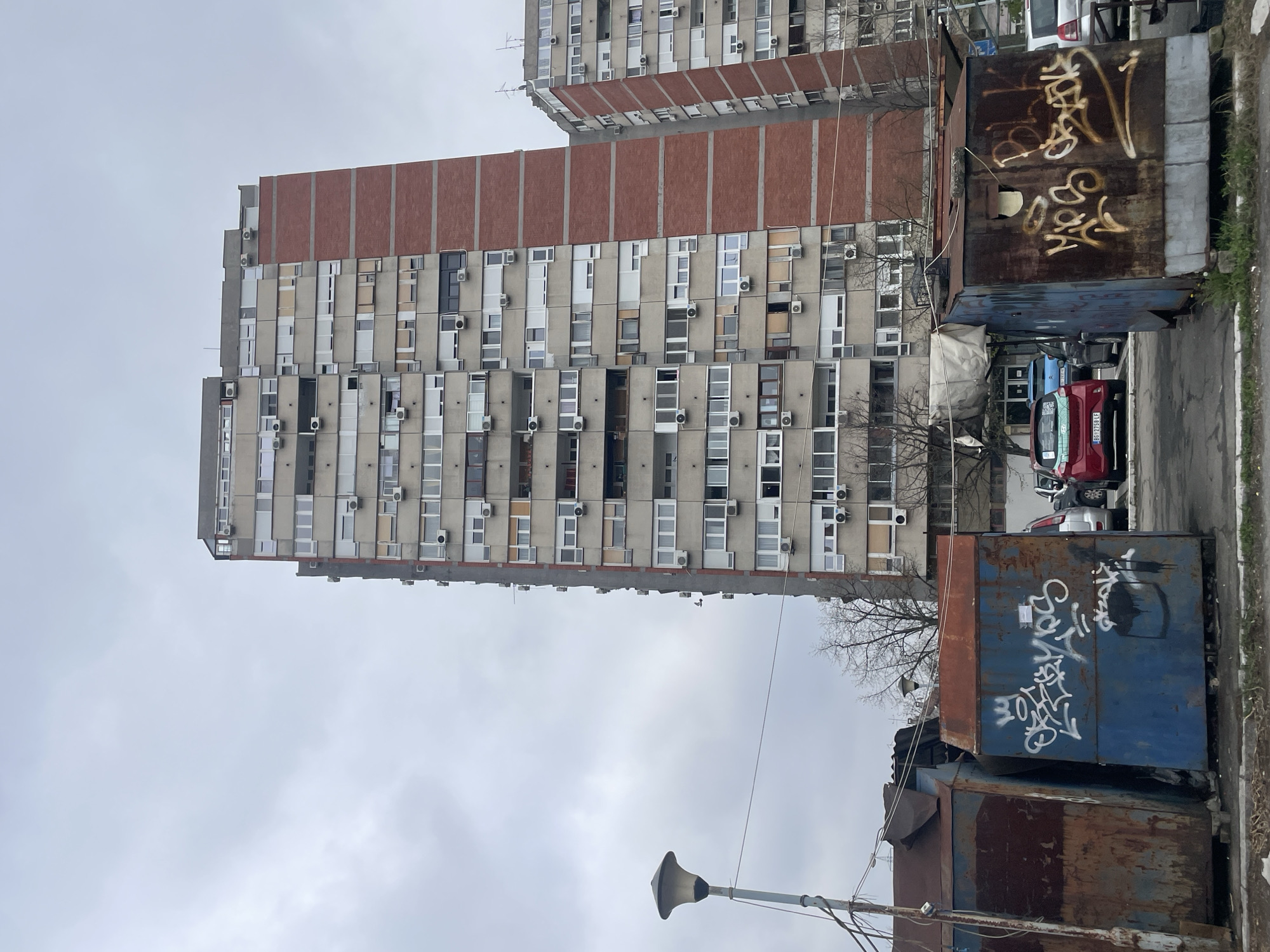 The image depicts a tall, multi-story residential building in Belgrade, Serbia. The building is constructed with a concrete frame and features rows of windows and air conditioning units. A section of the building has a brick facade. In the foreground, there are several large metal dumpsters covered in graffiti. A few parked cars, including a red one with license plates visible, are situated behind the dumpsters. The sky is overcast, suggesting a cloudy day. A street lamp is visible in the lower part of the image. The overall scene appears urban and slightly rundown.