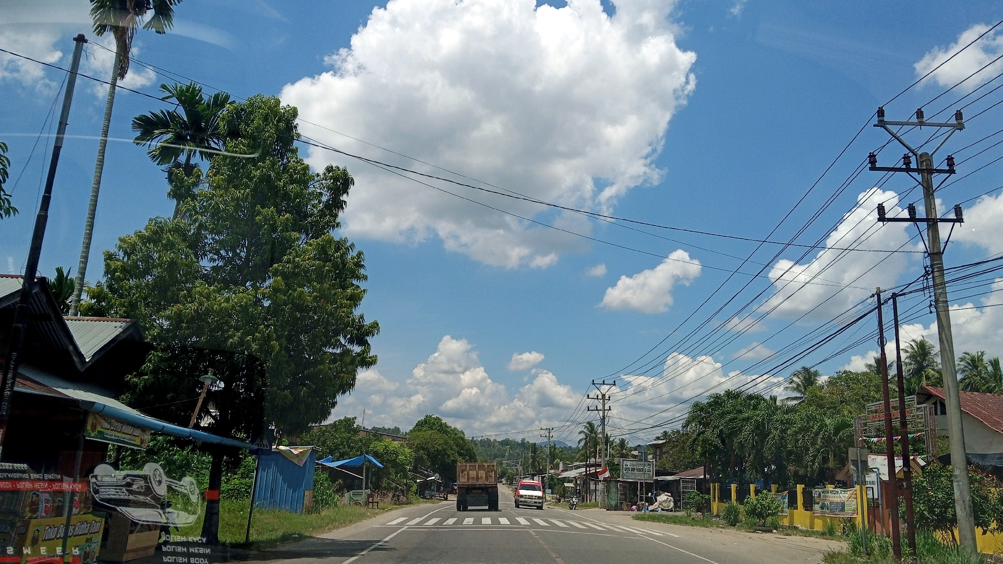 The image shows a street scene in the city of Beunyot, Indonesia. A two-lane asphalt road runs through the center of the frame, with a white dashed line marking the center. On the left side of the road, there are several small shops or stalls with corrugated metal roofs, one of which displays various items. A large green tree stands prominently near the shops. On the right side of the road, a utility pole with numerous wires is visible, along with more shops and houses. Palm trees and other lush green vegetation are scattered throughout the scene. The sky is a bright blue with scattered white cumulus clouds. Two trucks, one a dump truck and the other a smaller white truck, are driving away from the camera on the road. The overall impression is a typical street in a tropical, likely rural or semi-urban, Indonesian setting.