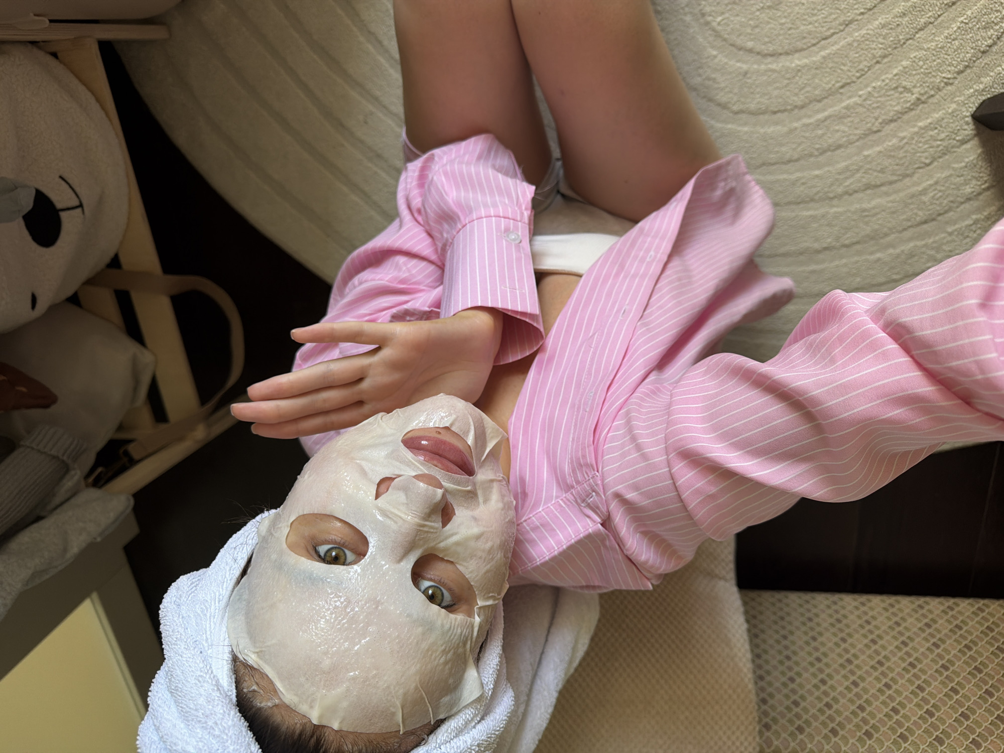 A woman is lying on a bed with her face covered in a sheet mask, a white towel wrapped around her head, and wearing a pink and white striped shirt. She is holding her hand up near her face. To the left of the woman is a stuffed animal on a wooden chair. The bed has a light-colored textured blanket and is next to a dark brown surface. The photo was taken in Khimki, Russia.